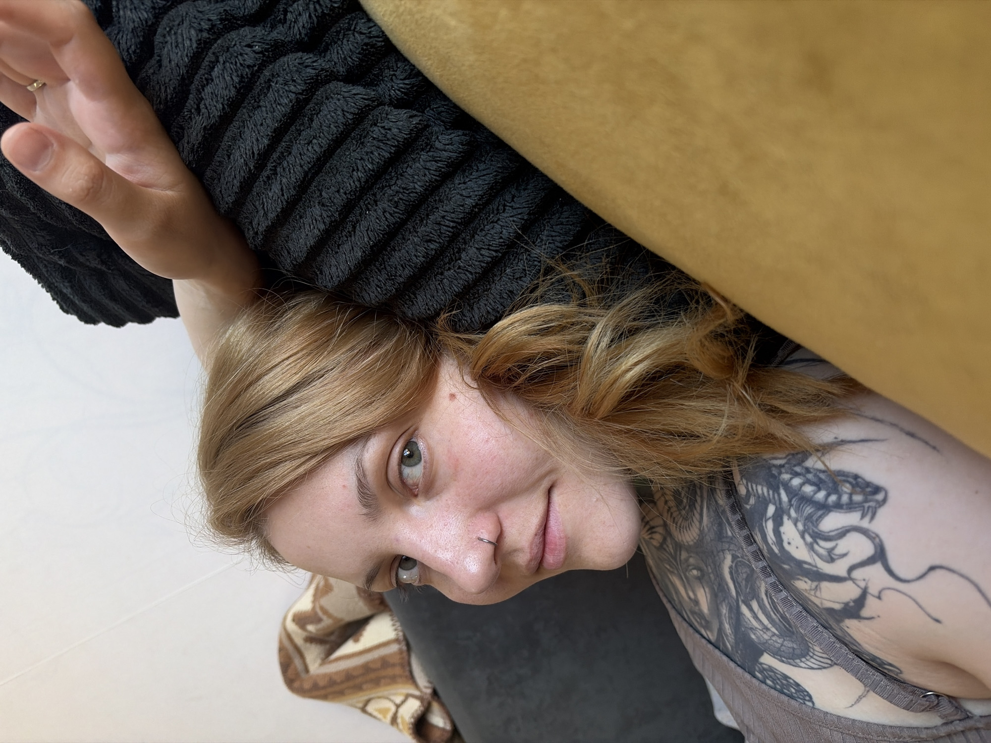 A person with light brown hair and a nose ring is lying down next to a black cushion and a yellow object. The person has a tattoo of a snake and a woman on their upper body. The background includes a white floor and a brown patterned object. The location is unknown, and there is no indication of Chaikovsky, Russia.