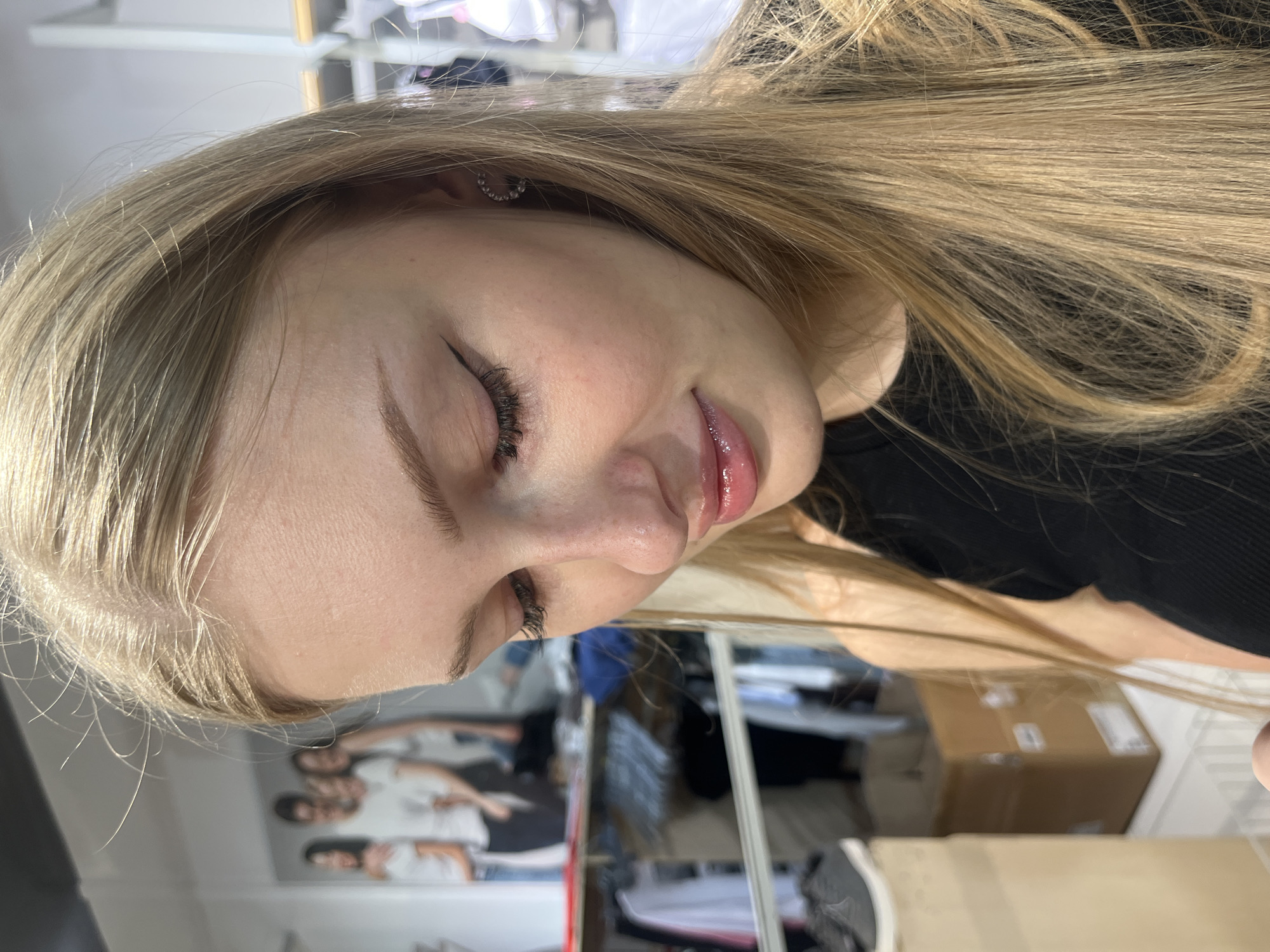 A young woman with long blonde hair is shown in a close-up, profile shot. She is looking down, and her eyes are closed. She is wearing a black top and has a small, circular earring with rhinestones in her left ear. Her lips appear glossy. In the background, there are clothing racks with garments, boxes, and a poster featuring a group of people. The environment appears to be a retail store. The location is not specified in the image.