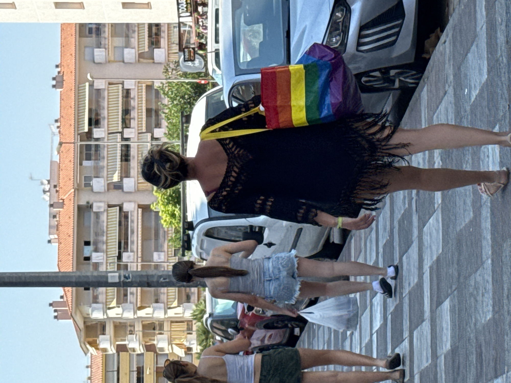 Three women are walking away from the camera on a sidewalk in Blanes, Spain. The woman in the foreground is wearing a black, fringed cover-up over a dark top and is carrying a rainbow-colored tote bag over her left shoulder. The middle woman is wearing a gray tank top and denim shorts, carrying a white plastic bag in her right hand. The woman on the left is wearing a white tank top and a green skirt. The background shows buildings with red-tiled roofs and apartment balconies, along with parked cars.