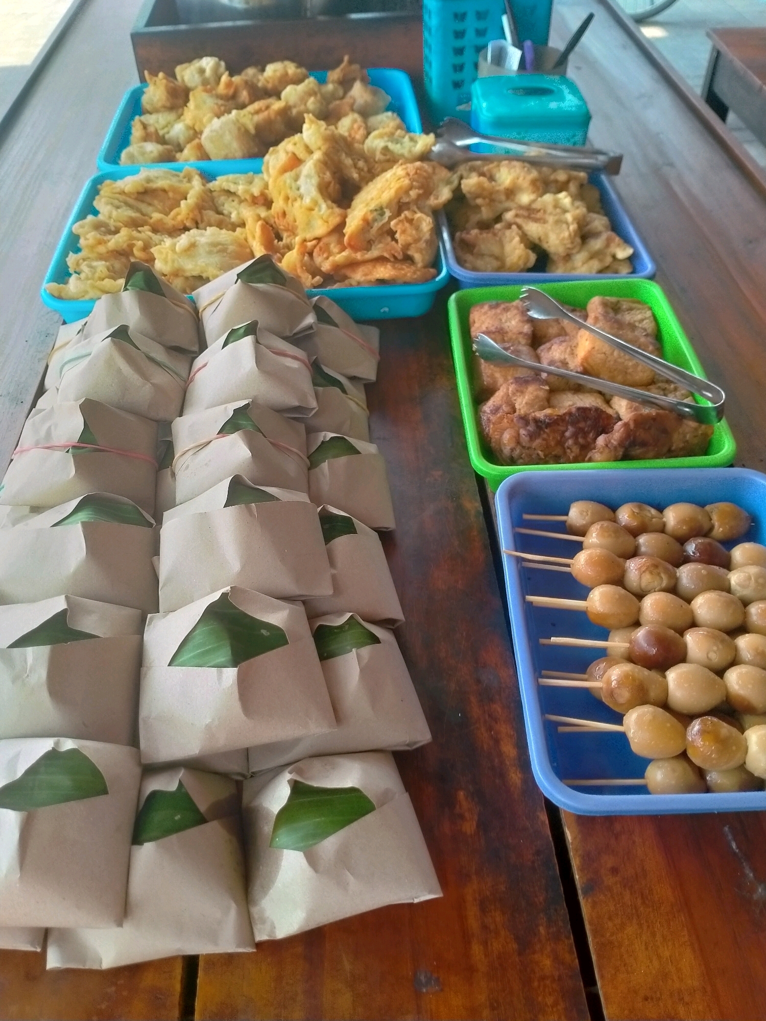 A close-up, eye-level shot captures a wooden table laden with various Indonesian food items, suggesting an outdoor market or food stall setting in Kasihan, Indonesia. In the foreground, a stack of rice packets, wrapped in brown paper and tied with rubber bands, are neatly arranged. Each packet has a piece of green leaf protruding from the top. To the right of the rice packets, a blue plastic tray holds skewers of quail eggs. The eggs are small, oval, and have a glossy, cooked appearance, with some showing a slightly darker brown hue. To the left of the quail eggs, a green plastic tray contains pieces of fried tofu or tempeh. Silver tongs are placed in the green tray, resting on the fried items. Further back, two blue plastic trays are filled with an assortment of fried foods, possibly vegetables or snacks, with a golden-brown, crispy texture. A dark wooden crate is partially visible in the upper left corner. In the background, a blue container with a patterned design and a smaller turquoise container sit on the table, along with a utensil. The overall scene is one of ready-to-eat food items displayed for sale.