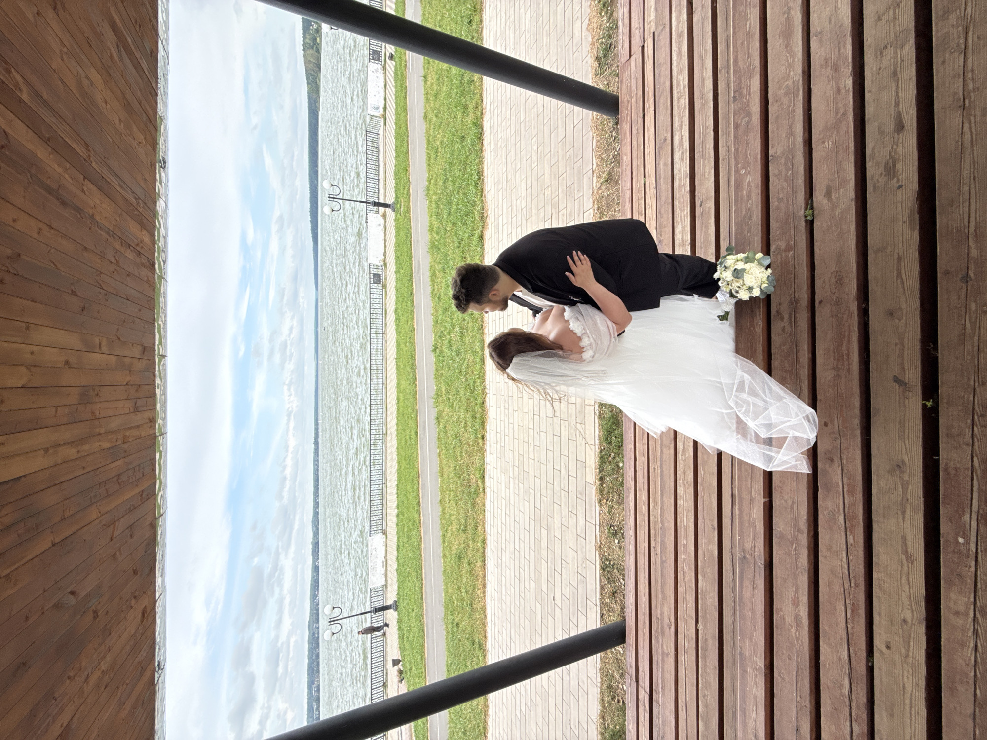An overhead shot captures a wedding couple standing on a wooden platform overlooking a lake. The bride, in a white gown and veil, is embraced by the groom, who wears a black suit. A bouquet of white flowers rests on the platform near their feet. The lake, under a cloudy sky, is expansive with a distant treeline and buildings along its shore. A paved walkway and a grassy area separate the platform from the lake. In the background, a person walks along the walkway, and decorative lampposts are visible. The scene is framed by the wooden beams of a roof overhead.
