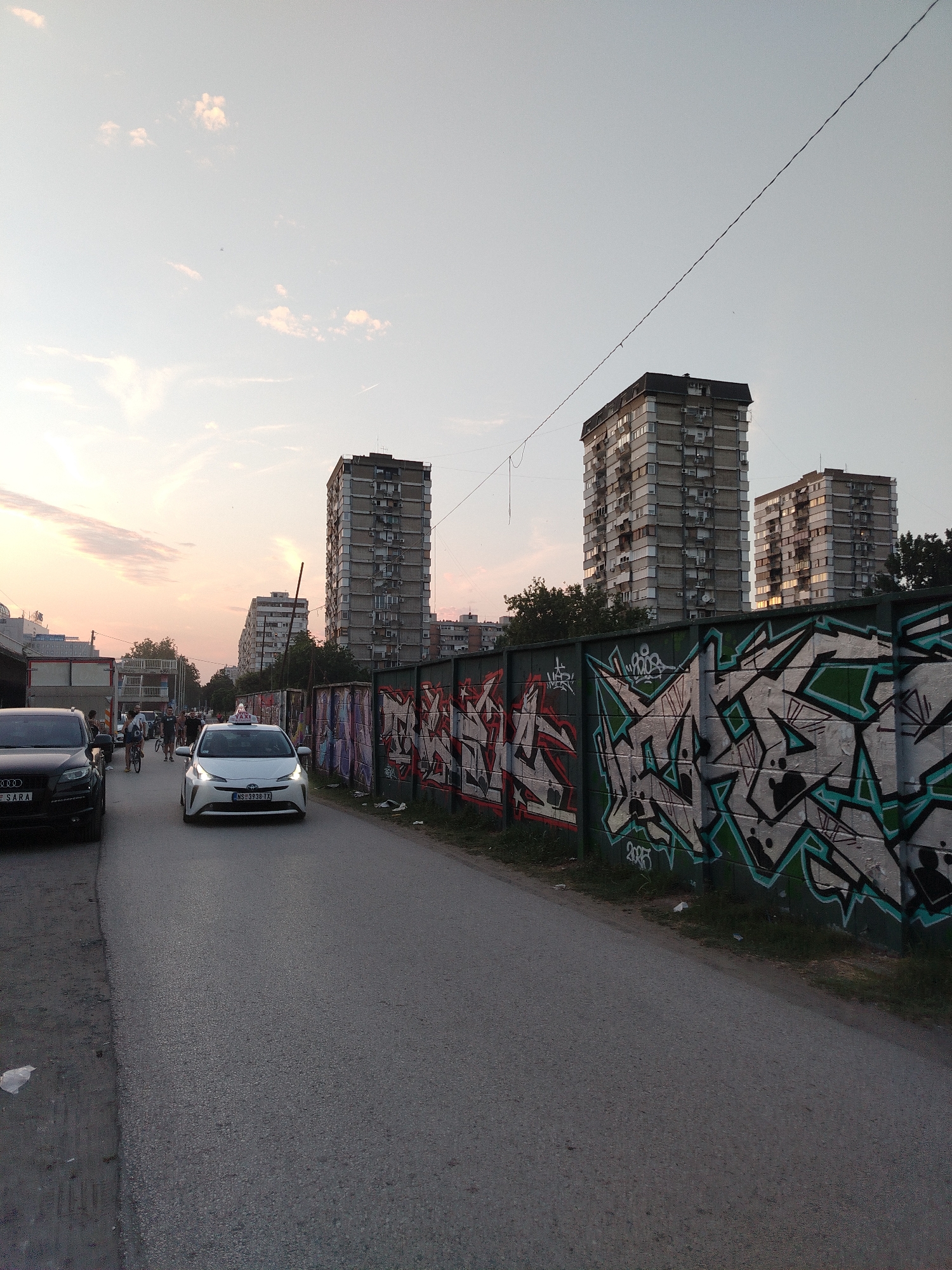 The image depicts a street scene in Novi Sad, Serbia, likely during the evening. In the foreground, a paved road runs diagonally from the bottom left towards the upper right. On the left side of the road, a black Audi SUV is parked. Further down the road, a white sedan, possibly a taxi, is in motion. Several people on bicycles are also visible on the road, indicating pedestrian or recreational activity.
The right side of the road is lined with a long, graffiti-covered wall. The graffiti features vibrant colors and intricate designs, characteristic of urban art. Behind the wall, trees and the facades of several tall, multi-story apartment buildings are visible. These buildings appear to be typical Soviet-era or post-Soviet residential blocks, with many windows and balconies. The sky is a muted orange and pink, suggesting a sunset or twilight atmosphere. The overall impression is one of an urban environment with elements of both everyday life and artistic expression.