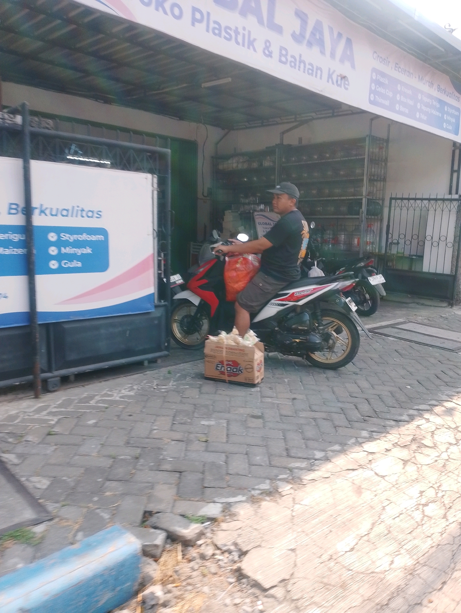 In Probolinggo, Indonesia, a man wearing a dark baseball cap and a black t-shirt with a graphic print is seated on a white and red motorcycle. He is wearing brown shorts and has a large orange bag behind him. The motorcycle has gold rims and a license plate that reads "L 2820 UJ". In front of the motorcycle, on the paved ground, is a cardboard box with a yellow and red logo that says "Enak". The man appears to be leaving or arriving at a storefront, which has a blue and white sign with text including "Berkualitas". The storefront has shelves filled with various items. The ground is paved with interlocking bricks, and in the foreground, a blue wooden plank is visible. The scene is lit by sunlight, creating shadows.