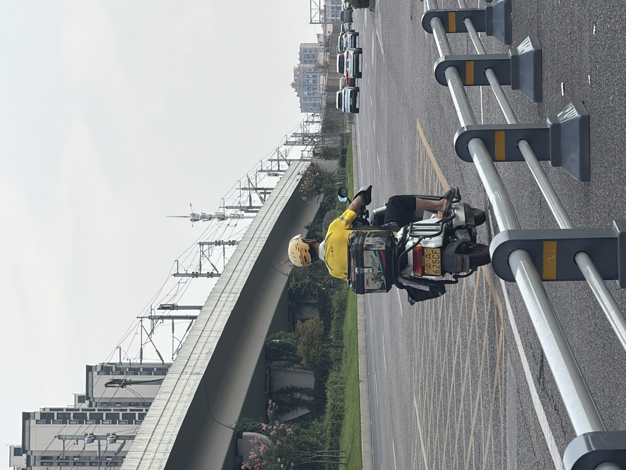 In Jiangjin, China, a delivery person wearing a yellow shirt and helmet rides a white electric scooter on a multi-lane road. The scooter has a large black storage box with colorful decals on the back. The scooter's license plate, partially visible, reads "A 625CF". In the background, a modern elevated overpass with train tracks runs parallel to the road, flanked by tall residential buildings on the left. On the right side of the road, several cars are driving, and further in the distance, more buildings are visible. The scene is bordered by metal guardrails in the foreground. The sky is overcast.