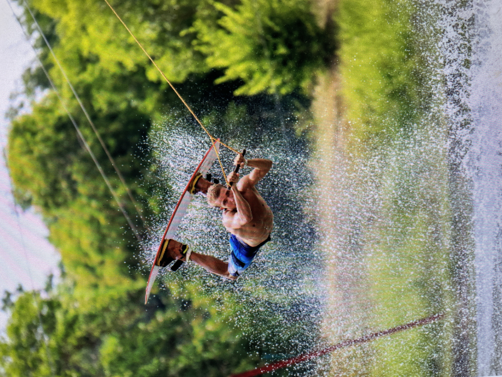A man is wakeboarding. He is shirtless, wearing blue swim trunks. He is in the air, holding onto a tow rope attached to a wakeboard. Water is spraying around him. Green trees are in the background. The image was taken in Bacong, Philippines.