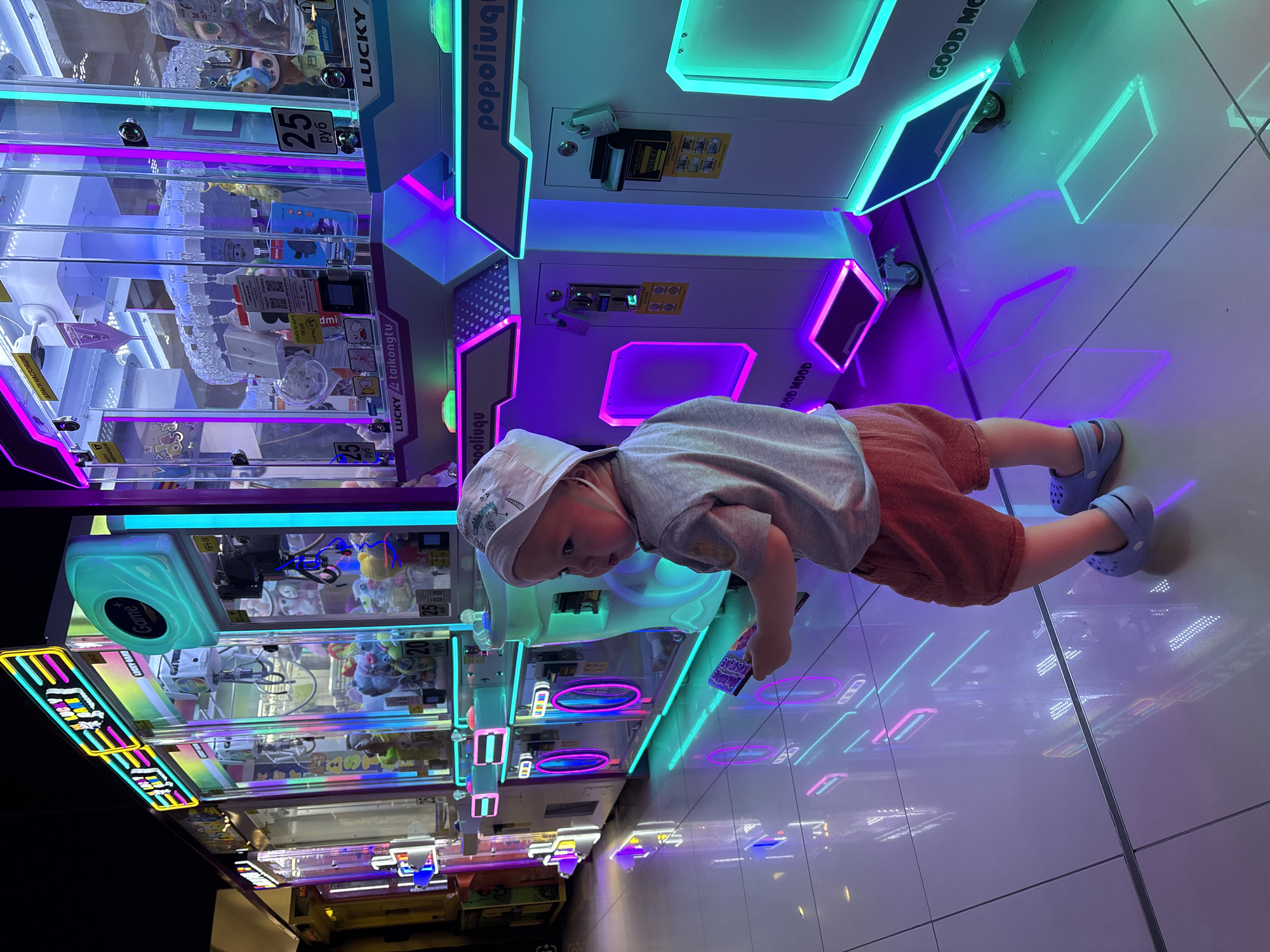 A young child wearing a light-colored bucket hat, a gray t-shirt, red shorts, and blue Crocs stands in an arcade. The child is facing away from the camera, looking towards a row of brightly lit claw machines. The machines are illuminated with neon pink, blue, and green lights, creating reflections on the glossy floor. The child holds a small purple object in their right hand. The background features multiple claw machines, some displaying prices in rubles and labels like "LUCKY TAİKONGTU" and "POPOLIUQU." The overall environment is an indoor arcade with a futuristic, vibrant aesthetic due to the intense lighting of the game machines. It is implied that this arcade is located in Naberezhnye Chelny, Russia.