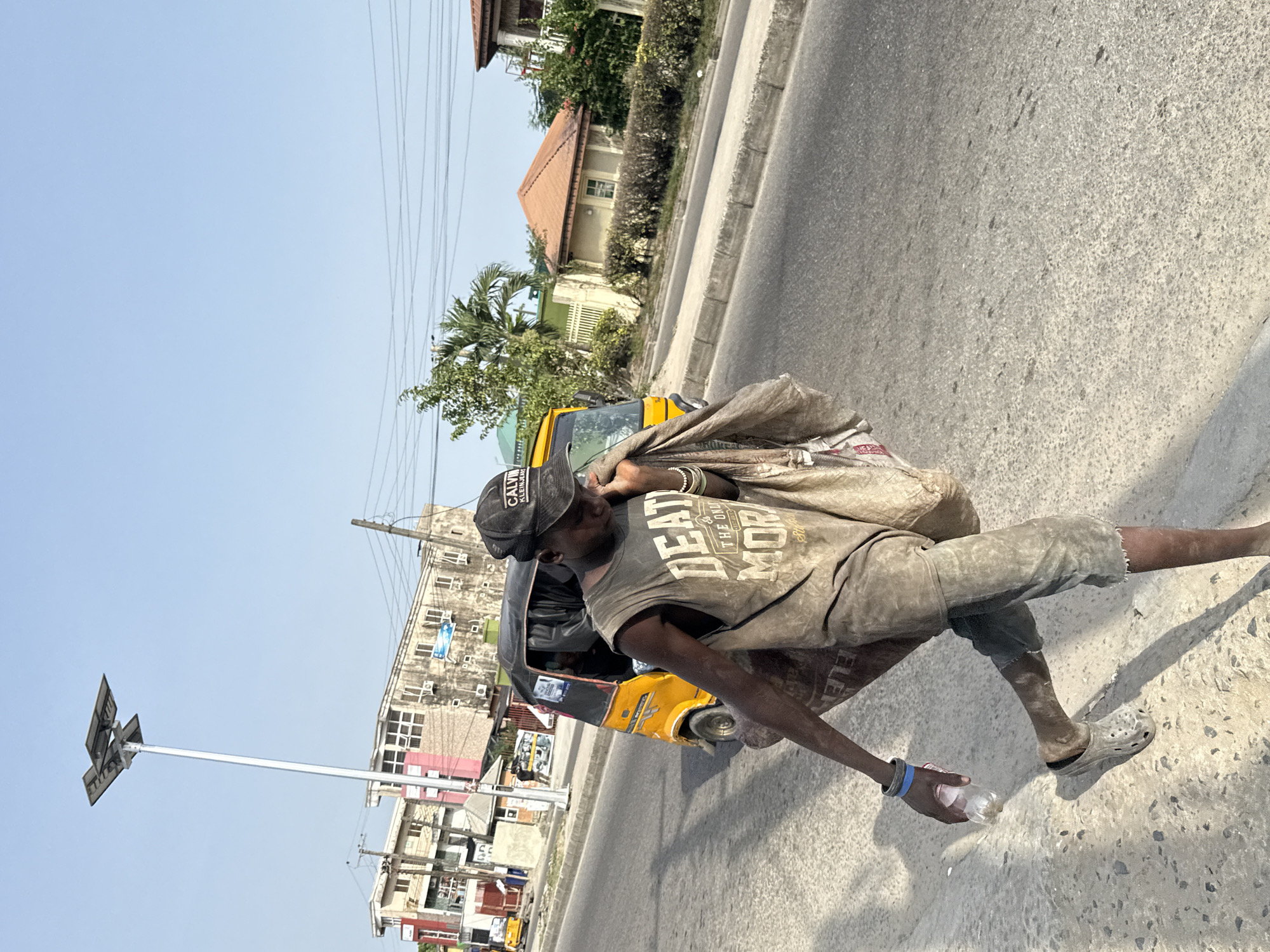 A mid-afternoon outdoor shot shows a man carrying a tan bag on his back, walking on a light-colored concrete sidewalk, wearing tattered brown clothes and a black cap, clutching an empty plastic water bottle in his hand. A three-wheeled yellow vehicle is on the road next to the sidewalk. Behind the vehicle, buildings in varying shades of tan and pink are visible. A tall, thin metal pole with four solar panels on top stands in the foreground. The photograph was taken in the city of Ado, Nigeria.