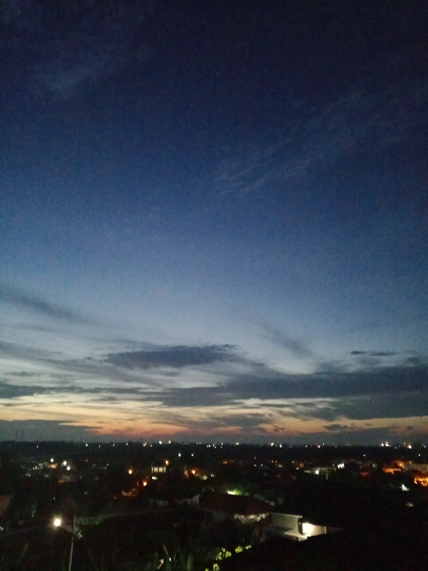 The image shows a twilight cityscape in Lhokseumawe, Indonesia. The sky transitions from dark blue above to a strip of faint orange near the horizon, partially obscured by low clouds. The city below is dark, with scattered lights indicating buildings and streets. The lower portion of the frame is filled with the dark outlines of trees and rooftops. A single street lamp is visible on the left. There are no people visible.