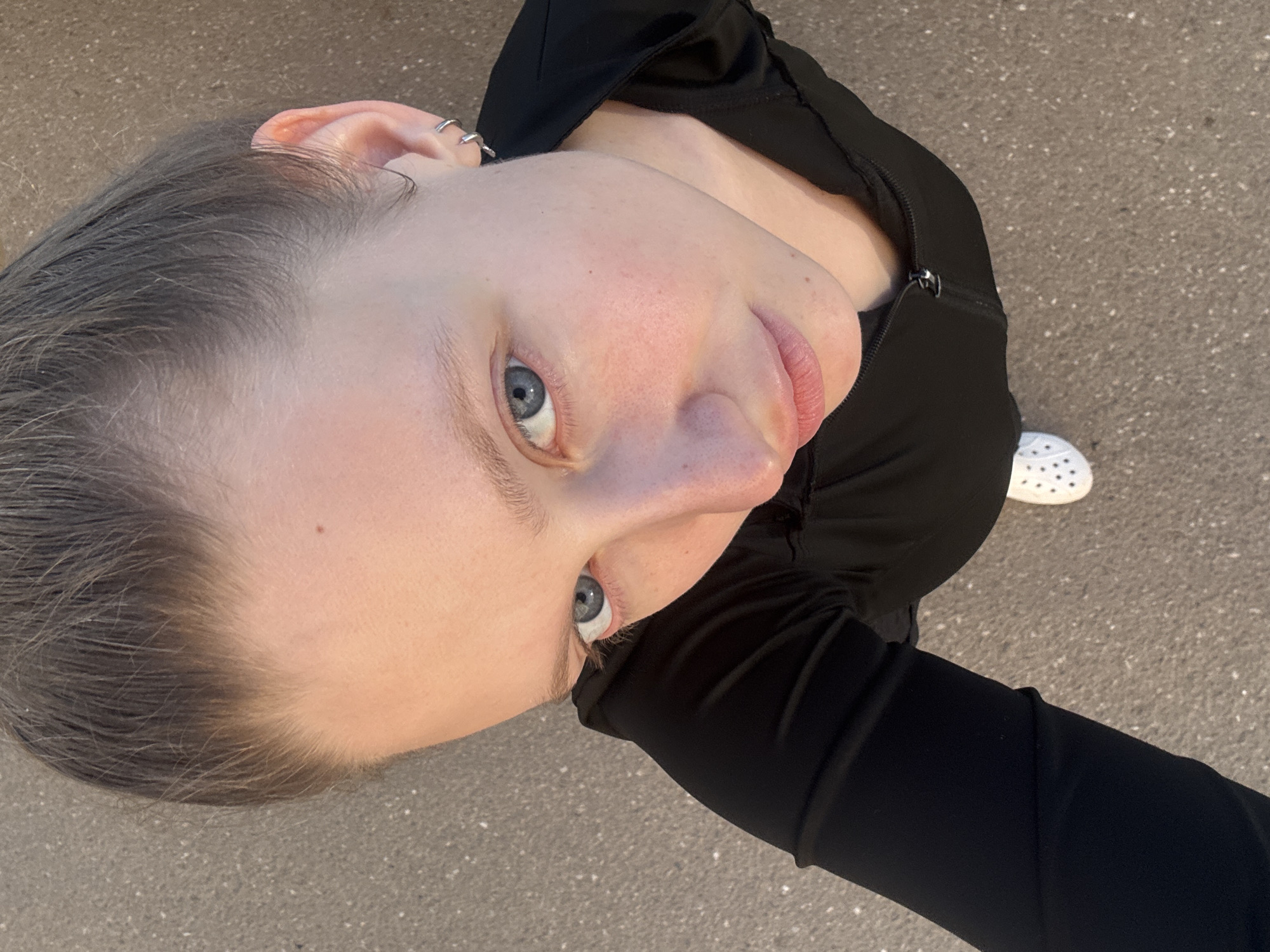 An overhead, low-angle shot captures a young adult looking directly up at the camera. The subject has fair skin, light brown or greyish hair, and striking blue eyes. Their lips are slightly parted. The visible ear features multiple silver hoop earrings. They are wearing a black long-sleeved top or jacket, possibly athletic wear, with a zipper visible at the neckline.
The photograph is taken outdoors, with a textured, greyish-brown pavement forming the background. The lighting is bright and natural, suggesting it is daytime, possibly sunny given the clarity. Part of a white Croc-style shoe with characteristic holes is visible in the lower right portion of the frame. The overall impression is a casual, spontaneous outdoor portrait. There is no visible text in the image. The location context is Izhevsk, Russia.