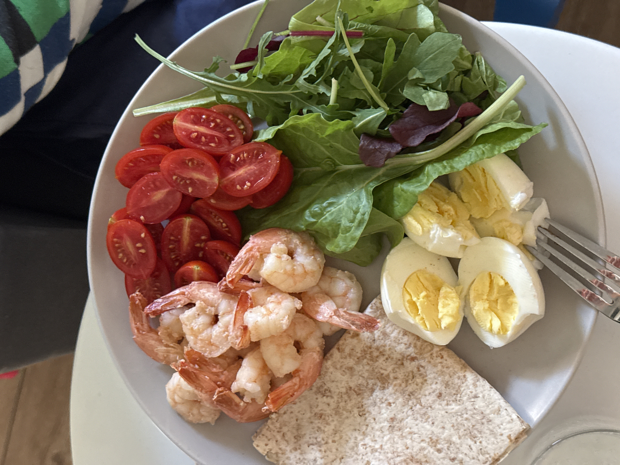 The image shows a close-up of a healthy meal served on a light gray plate. The meal consists of several cooked shrimp, halved cherry tomatoes, a portion of a whole wheat flatbread, a salad composed of mixed greens including arugula and red lettuce, and two hard-boiled eggs, each cut in half to reveal the yolk and white. A silver fork rests on the bottom right of the plate. The background is blurred, but suggests an indoor setting, possibly a table with a light-colored surface and a dark object, possibly furniture, behind it. The lighting appears to be natural, coming from above and to the left. There are no people or discernible urban environmental cues to indicate a location in Tel-Aviv, Israel.