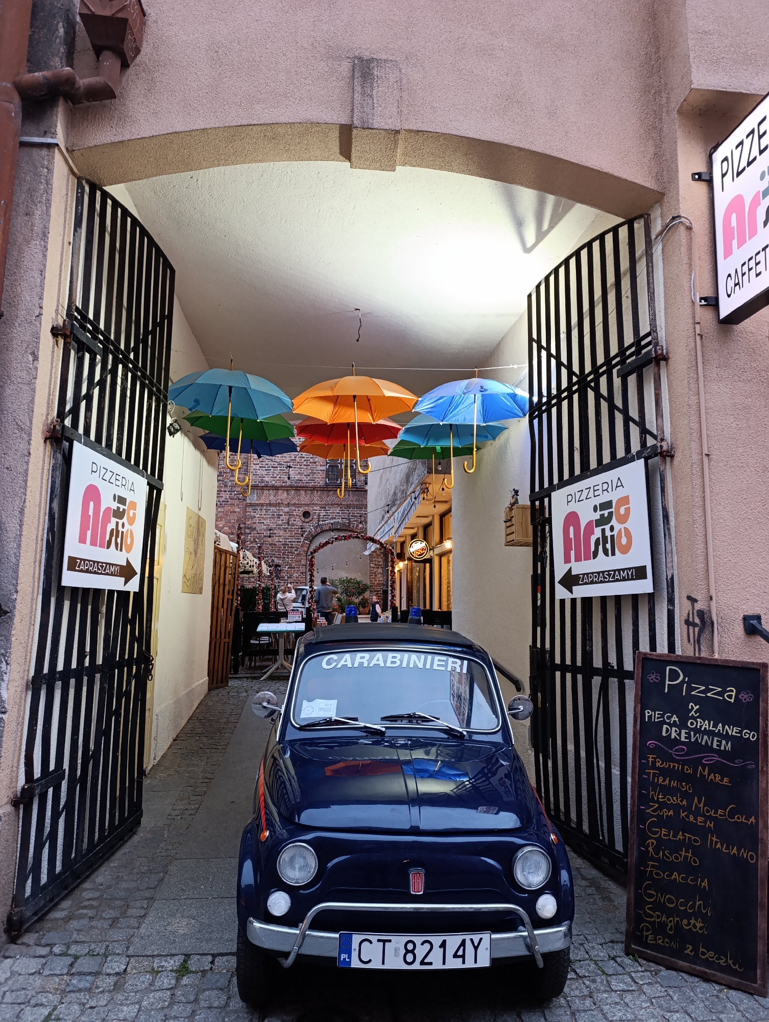 This media file captures a narrow, cobblestone alleyway in what appears to be a historic European city, likely Toruń, Poland. The setting is characterized by weathered brick buildings and an arched entryway draped with colorful umbrellas, creating a vibrant and inviting atmosphere.
Dominating the foreground is a vintage, dark blue Fiat 500 with "CARABINIERI" written across its windshield, suggesting a playful or thematic decoration. The car's license plate, "CT 8214Y," with "PL" indicating Poland, confirms the location.
To the left and right of the entryway, signs for "PIZZERIA Arti" with the Polish word "ZAPRASZAMY!" (We invite you!) are prominently displayed, indicating the presence of an Italian restaurant. A blackboard menu to the right lists various Italian food items, including pizzas, "Frutti di Mare," "Tiramisu," "Gnocchi," and "Spaghetti," as well as drinks like "Peroni."
In the background, through the archway, more seating areas with tables and chairs are visible, along with people enjoying a meal or gathering, suggesting a lively evening ambiance. The lighting appears to be artificial, hinting at dusk or evening. The overall impression is one of a charming, bustling dining district with a strong Italian theme.
