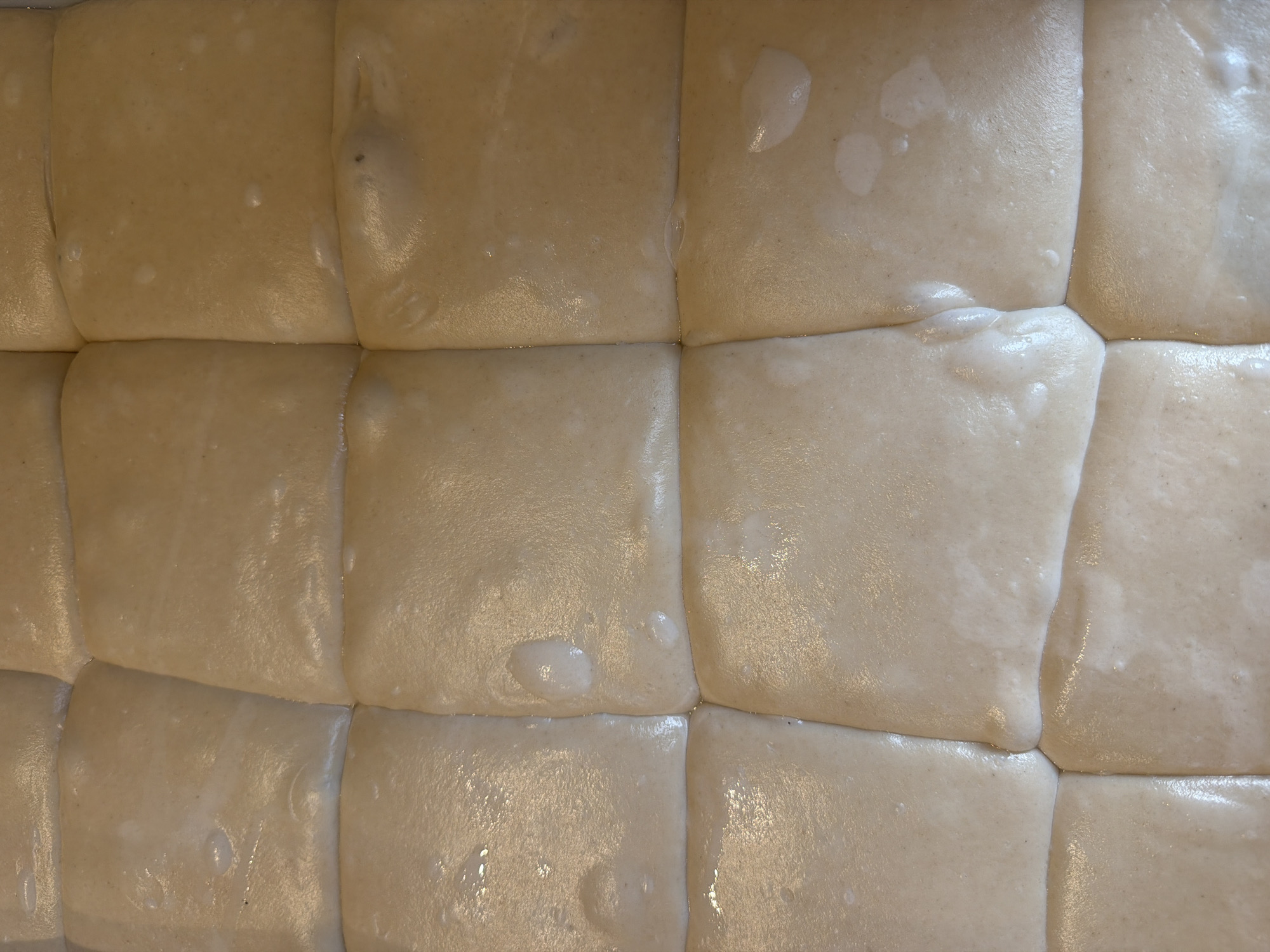 This is a close-up, top-down shot of a baking pan filled with twelve portions of dough, arranged in a 3x4 grid. The dough is a light tan color and appears to be raw and unbaked. Some portions have small bubbles or slight indentations on their surfaces. The dough has been scored or cut into individual squares, and there are visible lines separating each portion. The pan is not visible, as the shot is entirely focused on the dough. There are no people, animals, or other objects present in the image. The environment is a plain, indoor setting, suitable for food preparation.