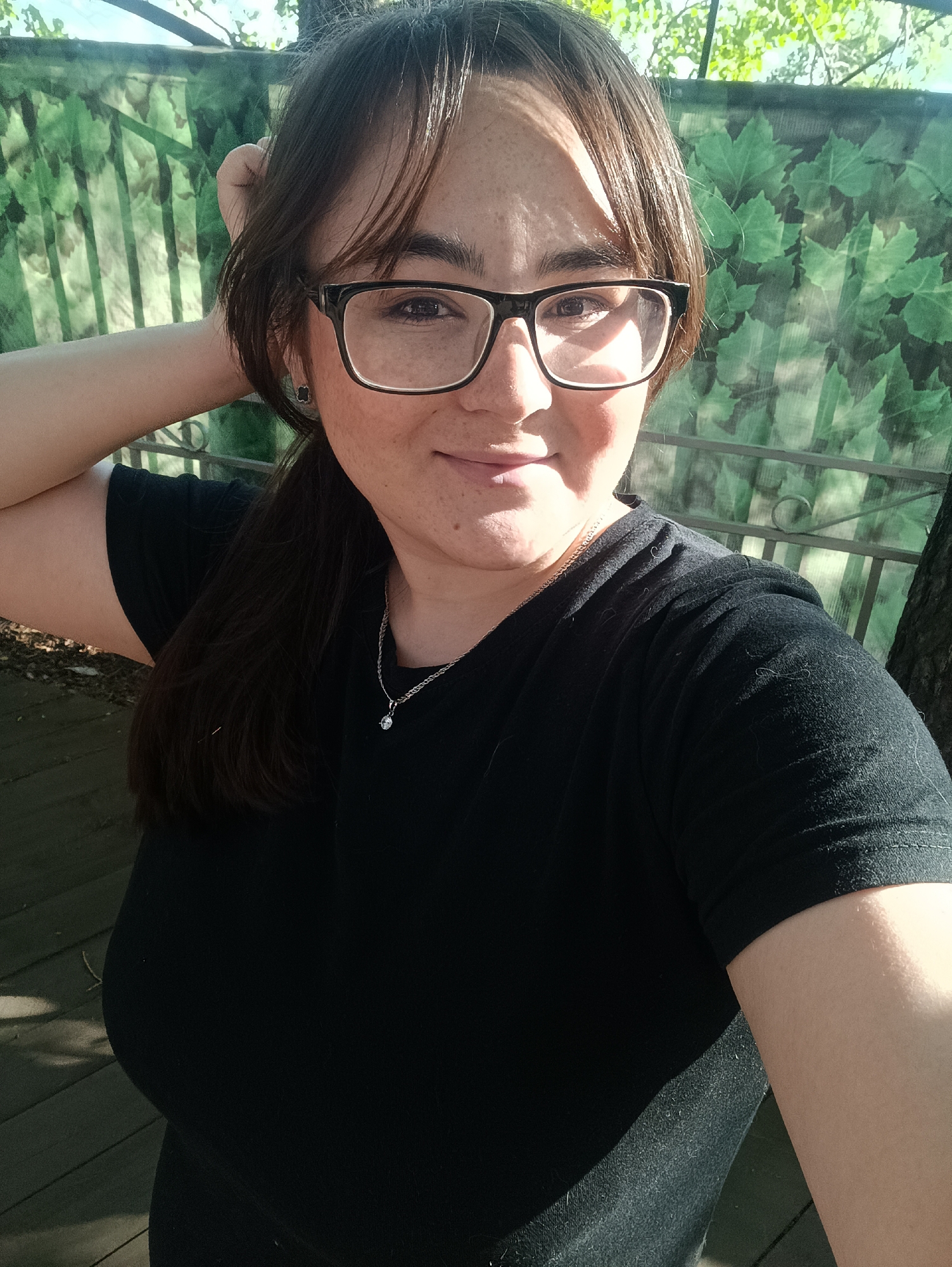 The image is a selfie of a woman with medium-length brown hair and glasses, wearing a black t-shirt and a silver necklace with a small pendant. She is standing outdoors, with a green leafy fence and a tree trunk in the background. The ground beneath her appears to be a wooden deck or boardwalk. Sunlight is casting shadows, indicating it is daytime. The overall impression is a casual outdoor portrait. The location is stated as Chaikovsky, Russia.