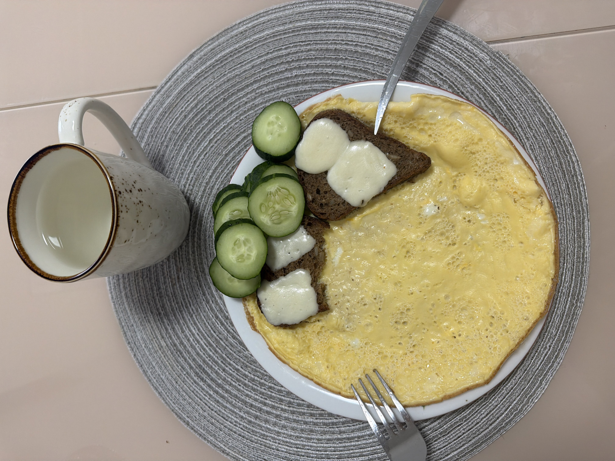 A top-down shot shows a white plate with a yellow omelet, topped with two slices of dark bread, each adorned with two irregular dollops of white cheese. To the left of the bread are several slices of cucumber. A fork and a knife are positioned on the plate. A white mug with a brown rim is placed to the left of the plate, resting on a gray, woven placemat. The scene is set on a light pink table. The location is not discernible from the image.