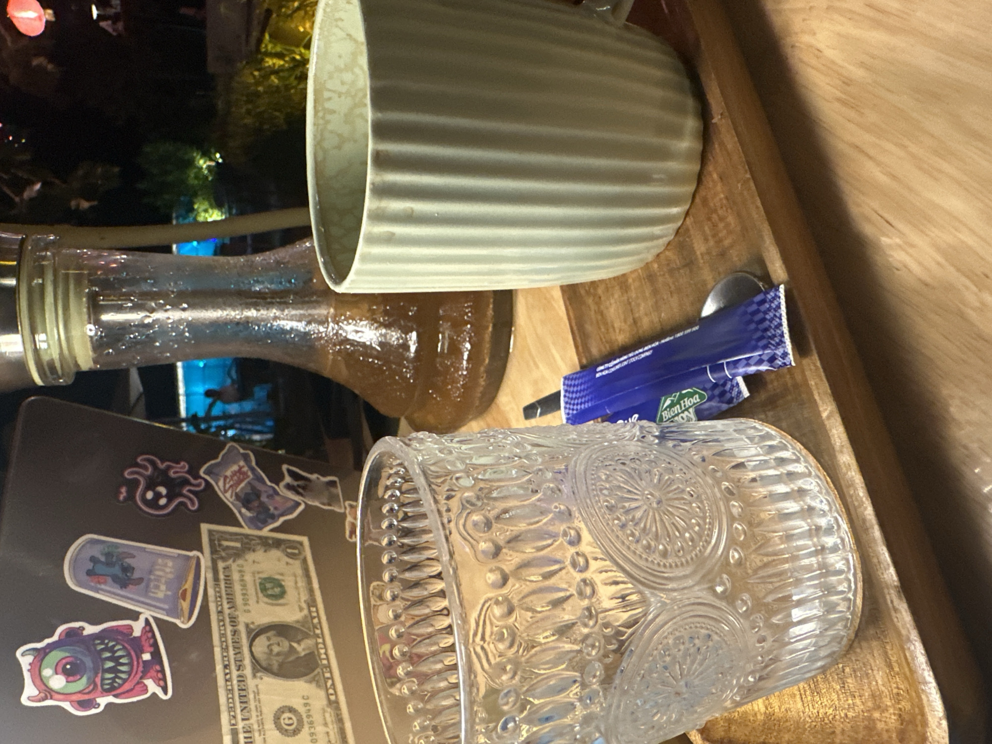This close-up shot features a table scene with several items. A large ceramic mug with horizontal ridges is near the top edge of the frame. Next to it is a glass bottle. A packet labeled "Bien Hoa" and a spoon are positioned on the wooden table. In the lower-left corner, a laptop covered with stickers (including a dollar bill sticker) is visible. The backdrop suggests an outdoor setting with vegetation and dim lighting, possibly taken in the ward of Bac Nha Trang, Vietnam, though there are no identifying landmarks confirming this. There are no people present.