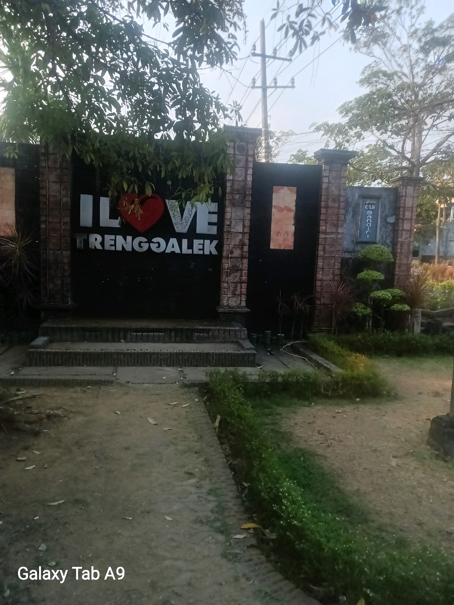 The image shows an outdoor scene with a brick wall and gate, adorned with a large sign that says "I LOVE TRENGGALEK." The sign is painted in white lettering, with a red heart symbol for the word "LOVE." The gate itself is black and flanked by brick pillars. To the right of the gate, there's a smaller sign that appears to have the text "CSR mandiri" in blue. In front of the gate, there are several steps leading up to it, and beyond that, a dirt path. To the right of the steps, there are some potted plants and small trees. The overall environment appears to be a public space, possibly a park or entrance to a facility, in Kelutan, Indonesia. The sky is visible in the background, with trees and power lines suggesting an urban or semi-urban setting. The image is taken from a low angle, focusing on the sign and the immediate surroundings. There are no people visible in the frame. The lighting suggests it is either early morning or late afternoon. The bottom left corner of the image displays "Galaxy Tab A9," indicating the device used to capture the photo.