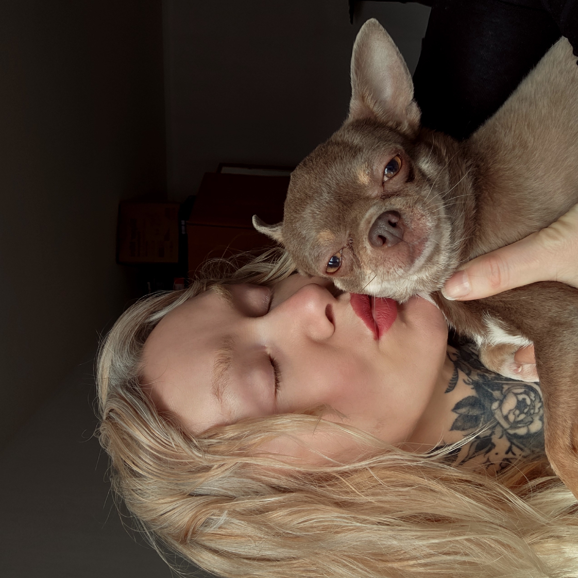 A woman with closed eyes and red lipstick is holding a light brown Chihuahua dog. The dog is resting its face on the woman's face. The woman has blonde hair and a tattoo on her neck. The background is dark. The location is not discernible, therefore no location will be provided.