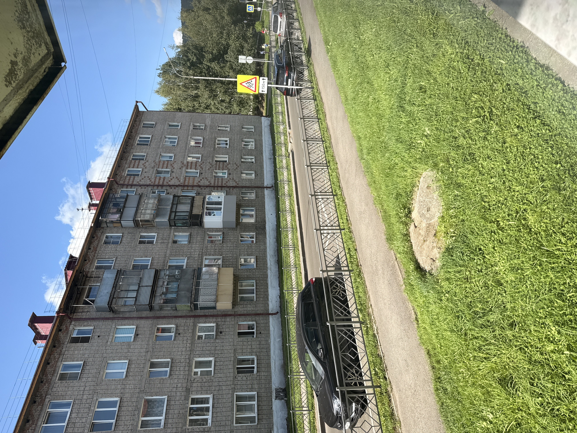 The image depicts a street scene in Mezhdurechensk, Russia, on a sunny day. A multi-story apartment building, constructed from gray brick, dominates the background. The building has numerous windows and balconies. In front of the building, a paved road is visible, with a sidewalk and a metal fence separating it from a grassy area. Two black cars are parked on the right side of the road, and a third vehicle is partially visible further down. A yellow triangular road sign with figures of children is posted on the right, indicating a pedestrian crossing approximately 150 meters ahead. Trees line the background, and the sky is blue with scattered clouds.