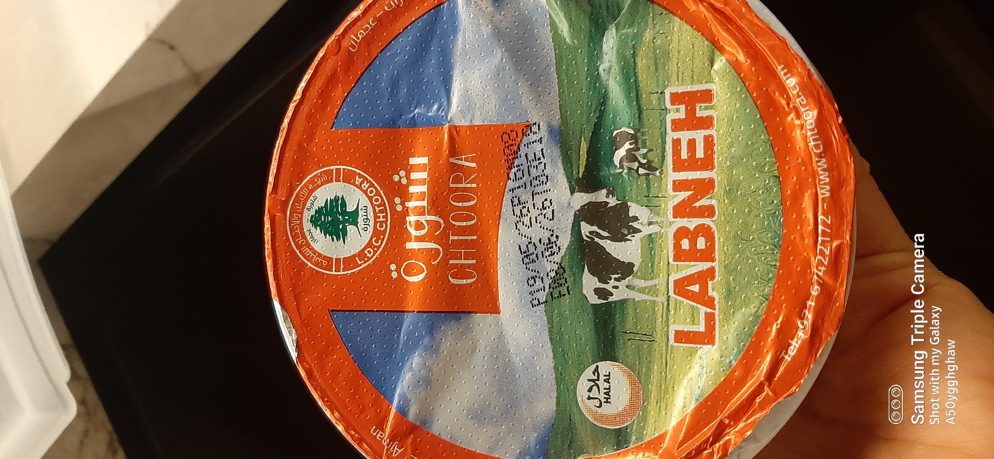 The image shows the top of a round food container, likely for cheese or yogurt, with an orange lid and a sticker on top. The sticker features a landscape with green hills and a blue sky, depicting cows in a field. There is text in Arabic and English. Prominently displayed is the brand name "CHTOORA" and the product type "LABNEH". A circular logo with a tree and Arabic text is visible at the top of the sticker, along with a "HALAL" certification. There are also printed date codes on the sticker. The lid itself has a textured orange surface. The container is partially held by a hand, and a portion of a Samsung Triple Camera watermark is visible at the bottom left, indicating the photo was taken with a Samsung Galaxy A50. The environment appears to be indoors with artificial lighting, and there is no specific indication of the city of Dubai, United Arab Emirates.