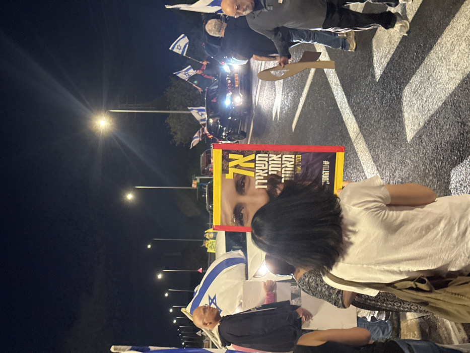 The image captures a nighttime gathering on a multi-lane road in Mishmar Hasharon, Israel. The scene is illuminated by numerous streetlights, casting distinct shadows on the asphalt.
In the foreground, a person with dark, shoulder-length hair is observed from behind, wearing a white t-shirt and carrying a light-colored shoulder bag on their right side. Directly in front of this individual, positioned centrally, is a large, rectangular sign with a prominent red and yellow border. The sign displays a close-up photographic image of a man's face with a beard and dark eyes. Hebrew text on the sign reads "אל תשאירו אותי מאחור" (Don't leave me behind) and includes the hashtag "#עדהחטוף" (Until the abductee).
To the left, a bald man wearing a dark hoodie is visible, looking towards the right side of the frame. Behind him, a large Israeli flag, predominantly white with a blue Star of David and blue stripes, is draped over a white vehicle or structure. Further to the right, an older bald man with glasses and a beard, wearing a dark top, holds a brown cardboard cutout shaped like a stylized missile or bomb.
In the mid-ground and background, several vehicles are present on the road, with their headlights illuminated. Some of these vehicles appear to have Israeli flags attached or displayed. Multiple Israeli flags are also held by individuals in the background. The sky above is dark, indicating it is nighttime.
