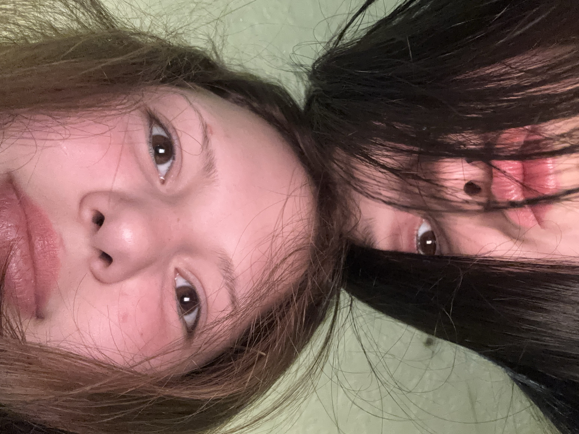 Two individuals are positioned closely together, their faces prominent in the frame. The individual on the left has light brown hair, while the individual on the right has dark, possibly black, hair. Both appear to have brown eyes. The background is a muted green color. The composition is an intimate, close-up shot. Location information is not discernible from the image.