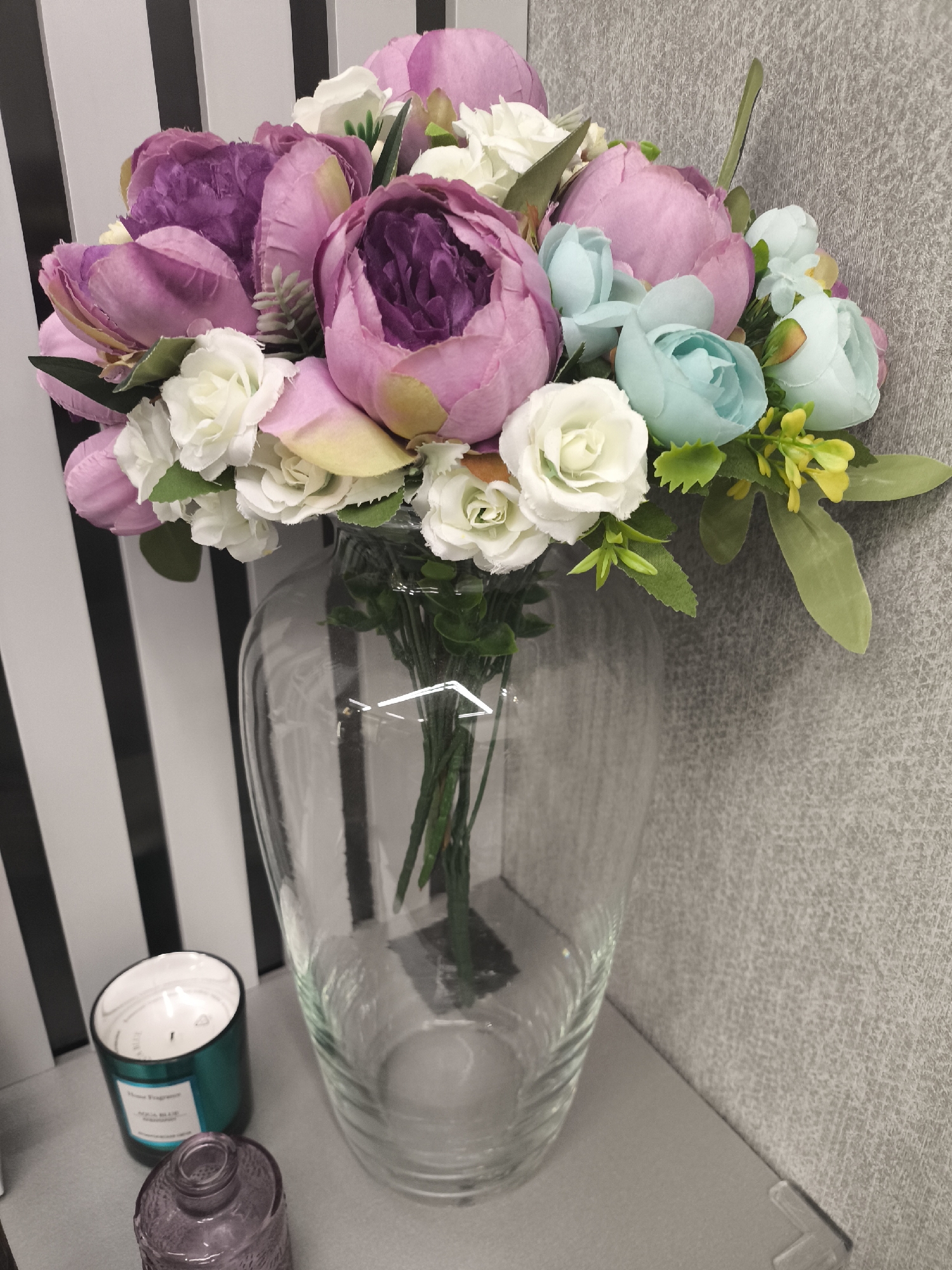 The image depicts a clear glass vase filled with artificial flowers of various colors including purple, pink, white, and blue. The vase sits on a gray surface, likely a shelf or tabletop, adjacent to a striped black and white background. A green candle and a small purple bottle are also visible on the surface. The setting appears to be an interior space, possibly a home or office. There is no indication of Moscow, Russia.