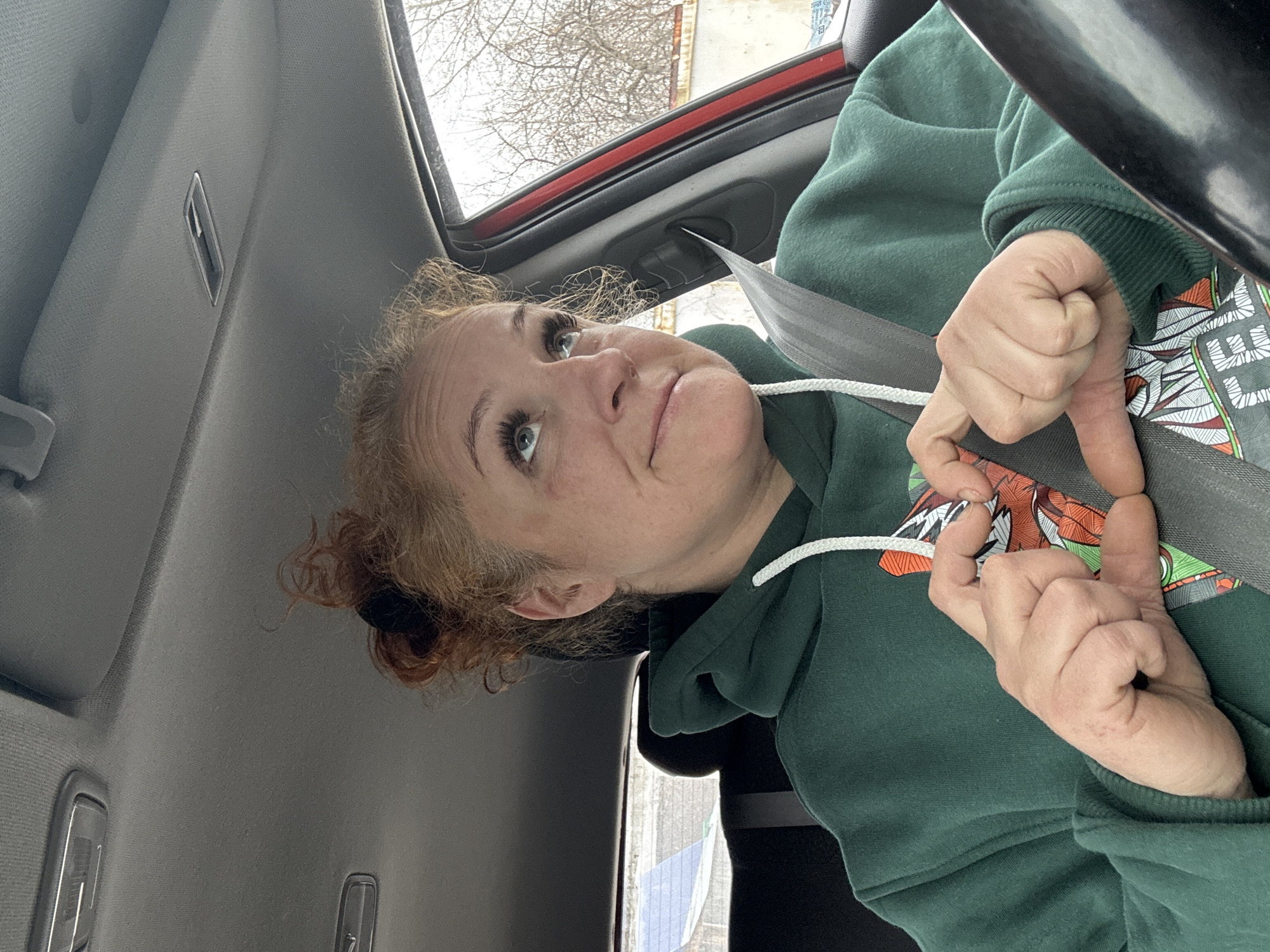 A fair-skinned, red-haired woman wearing a green hooded sweatshirt is seated in what appears to be the passenger seat of a car, indicated by the presence of a seatbelt. The car's interior is visible, including the ceiling and a red frame, likely the sunroof. Outside the car window, bare trees are visible, suggesting it may be winter or early spring. The specific location of Chaikovsky, Russia cannot be determined from this image.