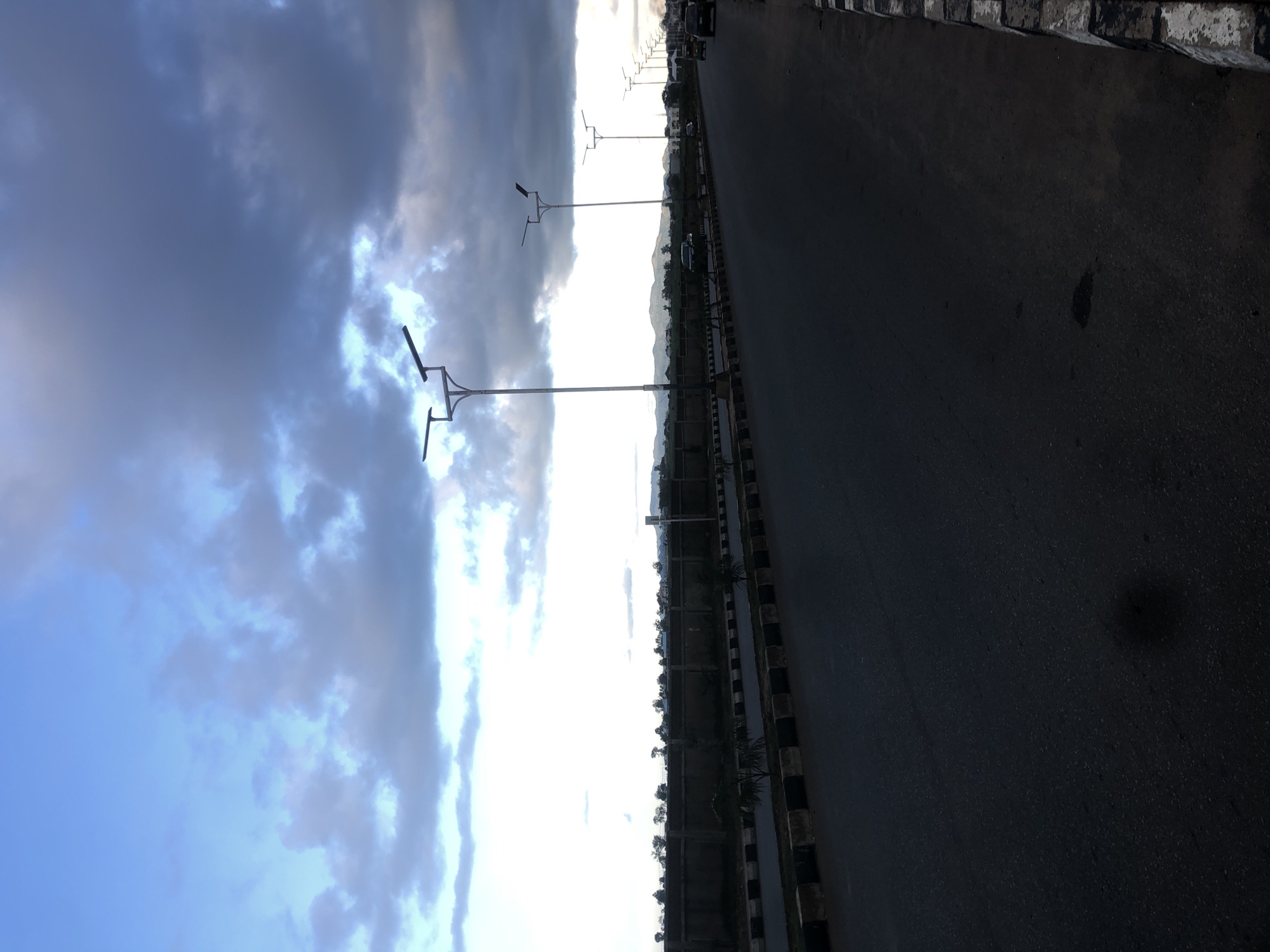 The image depicts a street scene in Barakin Lerawi, Nigeria, under a dramatic sky with dark, heavy clouds interspersed with patches of bright sky. A paved road stretches into the distance, bordered by a black and white striped kerb. On the left side of the road, a concrete wall runs along the edge, with small, sparsely planted trees and shrubs visible in front of it. Several modern streetlights with two lamps each are spaced along the right side of the road. In the background, a range of hills or mountains is visible. A few vehicles, including a black car and a white van, are present on the road. The overall impression is that of an urban or suburban area on the outskirts of the city, likely experiencing approaching inclement weather.