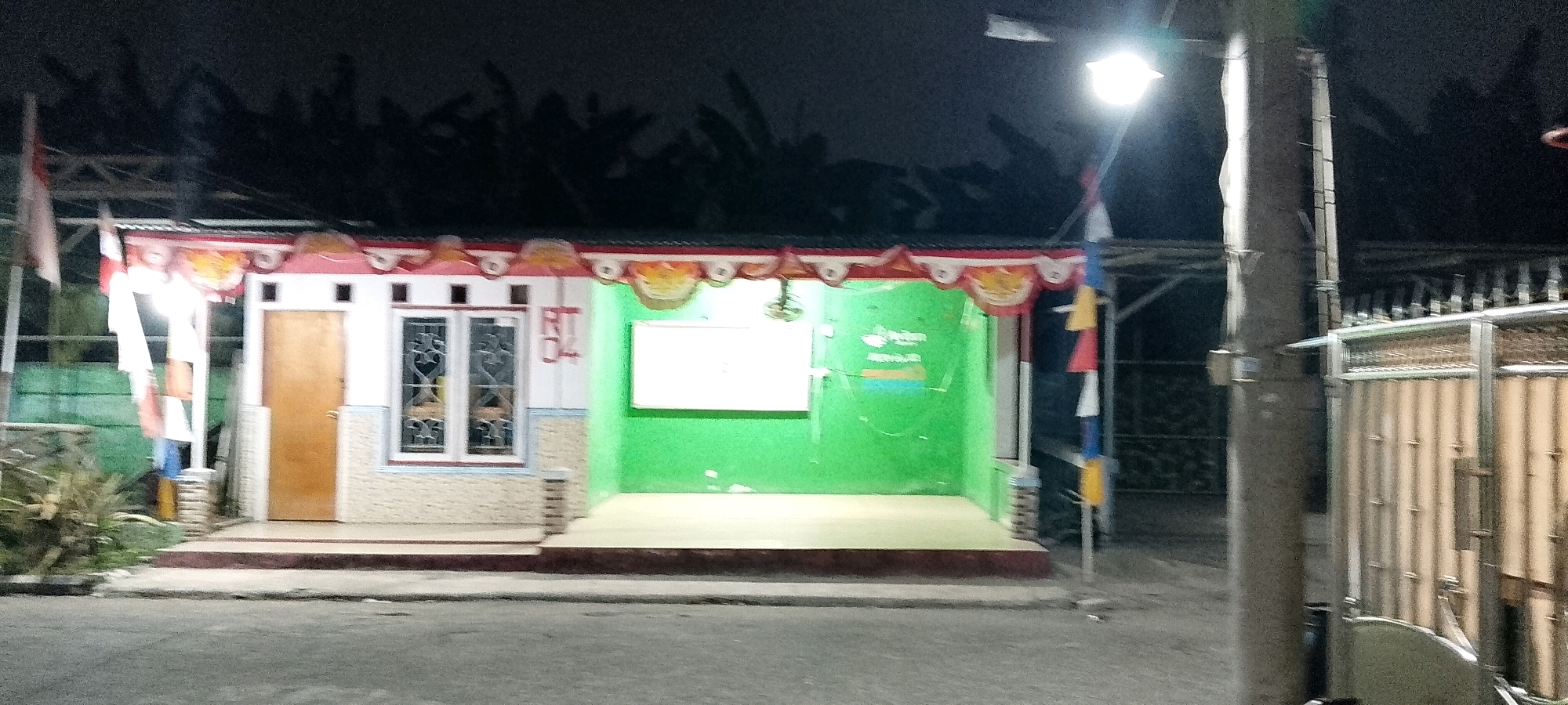 The image shows a small building at night, possibly a community post or small business, with a light pole and a fence in the foreground. The building has a white facade with a wooden door and a window with decorative grills. The roof is covered with corrugated metal and decorated with red and white trim and colorful fabric scallops. There is a green wall on the right side of the building, with a white rectangular board mounted on it. A small plant is visible to the left of the building. The scene appears to be in a residential area. The location is identified as Satriamekar, Indonesia.