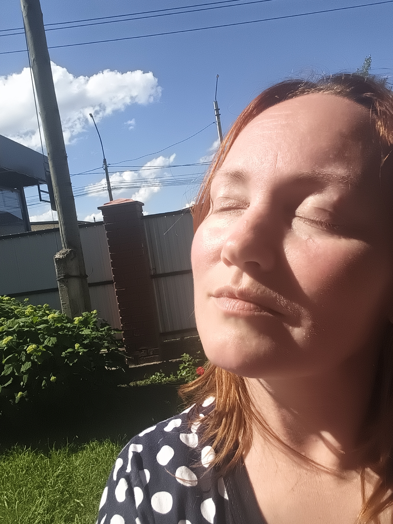 A person is captured in a close-up, side-profile shot, with their eyes closed and face turned towards the sun. The golden sunlight illuminates their face, highlighting its contours and the texture of their skin. Their hair, a reddish-brown, cascades around their face and shoulders. They are wearing a dark garment adorned with large white polka dots. The background reveals a green lawn, a lush bush with light green buds, a fence, a brick column, and a utility pole against a bright blue sky dotted with fluffy white clouds. The scene appears to be outdoors, possibly in a backyard or garden, during daylight hours. The location is identified as Chaikovsky, Russia.