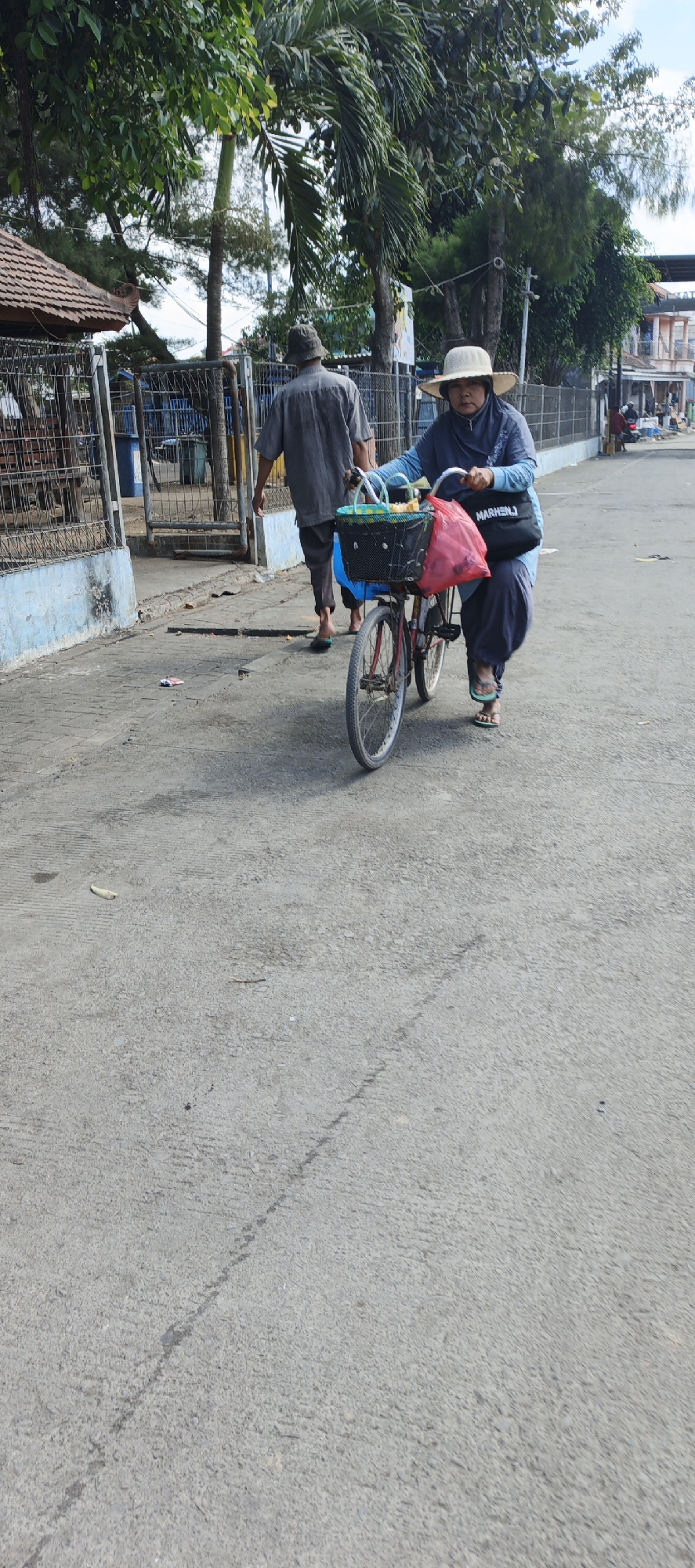 In the city of Puger, Indonesia, a woman wearing a wide-brimmed hat and a blue headscarf is pushing a bicycle. The bicycle has a black basket on the front containing a green and white checkered bag and a red plastic bag. A dark bag with "MARHEN.J" printed on it is slung over the woman's shoulder. She is wearing a blue top and dark pants, with light-colored sandals on her feet. Behind her, a person wearing a grey shirt and a dark hat walks away from the camera, with bare feet and blue pants. They are walking on a paved road with some debris scattered on the ground. To the left of the road is a fence and a building with a tiled roof. Trees and foliage are visible in the background. The scene appears to be outdoors, possibly in a marketplace or a pedestrian area.