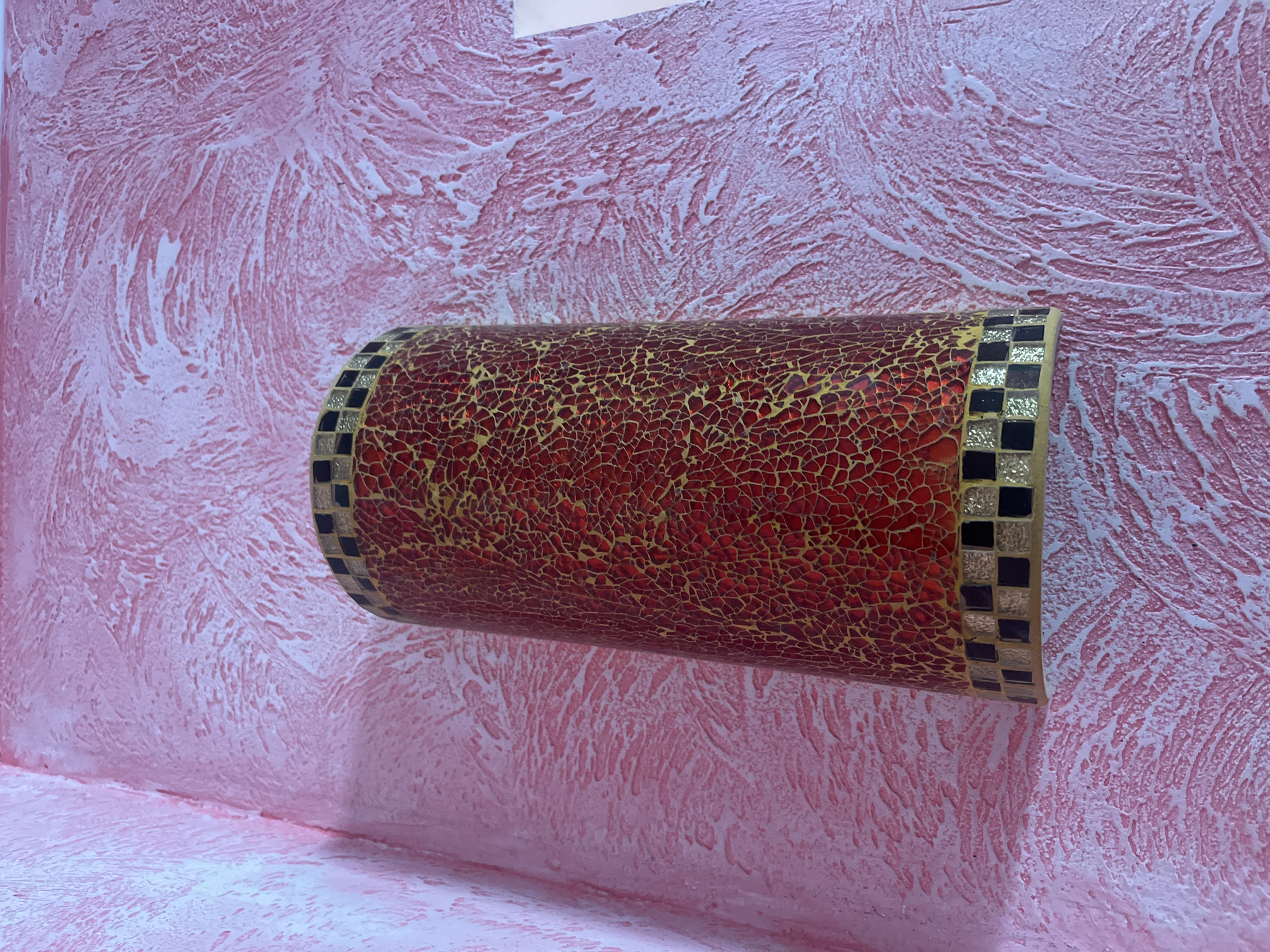 A cylindrical decorative light fixture, encrusted with a mosaic of red and gold cracked glass, is mounted on a textured pink wall in a corner. The mosaic pattern is intricate, resembling a network of fine lines. The top and bottom edges of the fixture are adorned with a border of alternating black and gold square tiles. The wall itself has a rough, stucco-like texture with visible brushstrokes or trowel marks, creating a visually dynamic surface. The lighting in the scene is soft, highlighting the textures of both the wall and the fixture. The location is specified as Tarnivtsi, Ukraine.