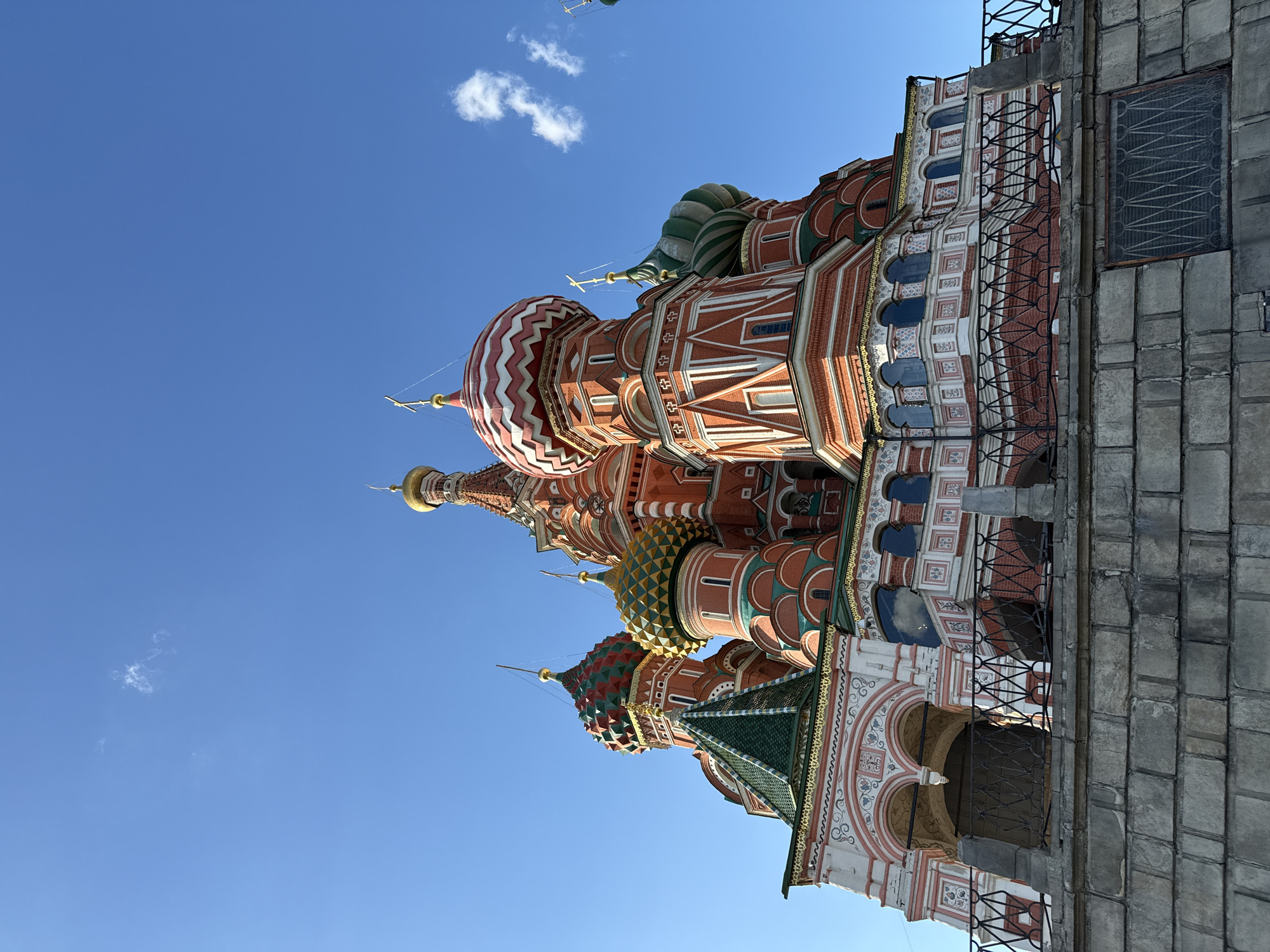 The image is a low-angle, upward-facing shot of St. Basil's Cathedral in Moscow, Russia, against a clear blue sky with a few wispy clouds. The cathedral's iconic, colorful onion domes are prominently featured. The building's facade is detailed with intricate brickwork, patterns, and decorative elements. The right side of the frame shows a section of the adjacent structure, constructed of gray stones, with a small window covered with a grill.