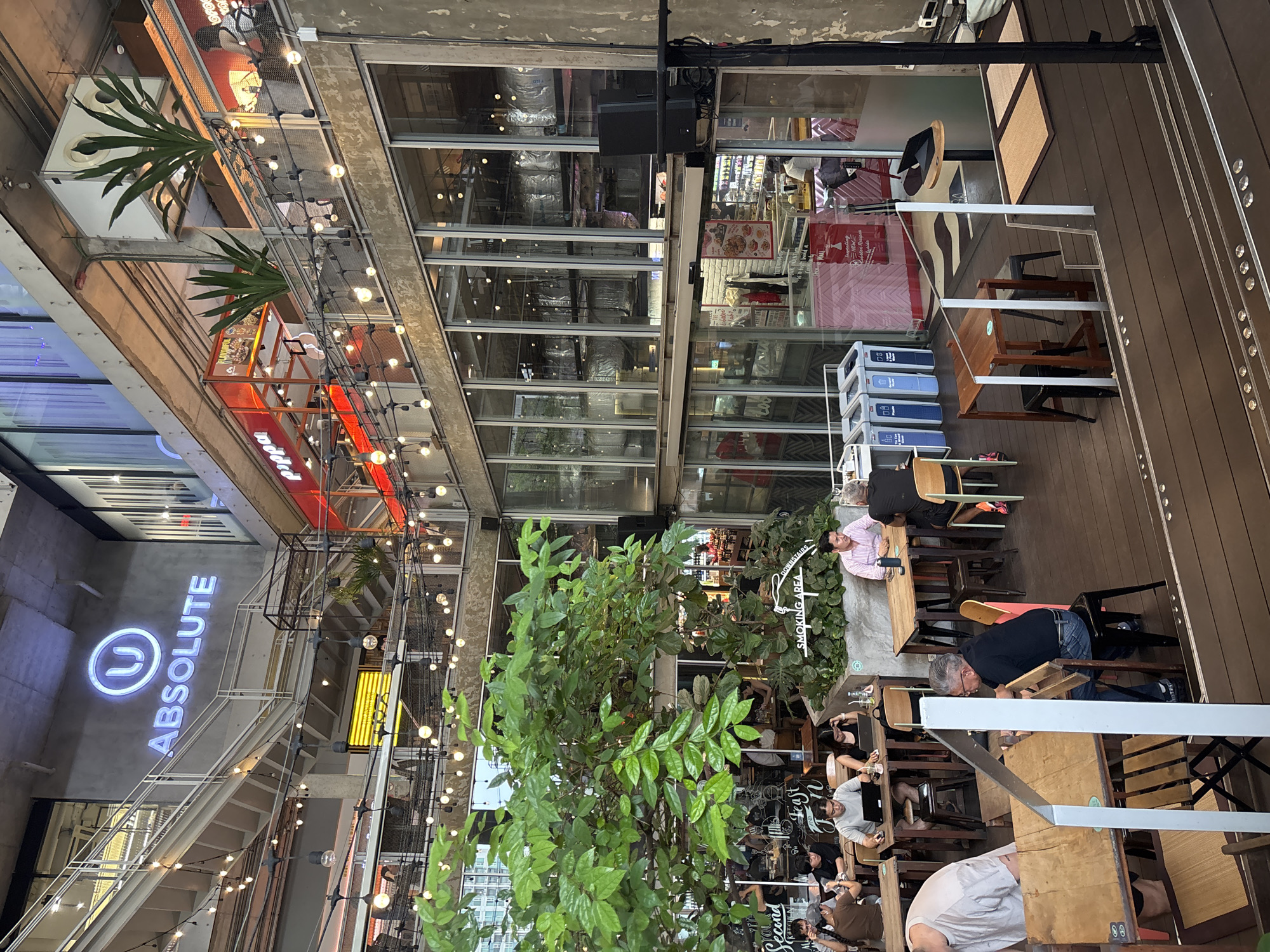 The image shows an outdoor dining area in a modern urban setting, potentially in Bangkok, Thailand. The area features a mix of restaurants and shops visible through glass walls. Diners are seated at wooden tables with metal chairs. Above, string lights are strung across the open space, and greenery is incorporated into the design. A sign for "Absolute" is visible, along with signs for restaurants like "Pippa" and "Second Kitchen", with the "DOWNSTAIRS" section noted as a "SMOKING AREA". Some individuals are dining while others pass by. The overall impression is of a bustling, contemporary outdoor dining and retail space.