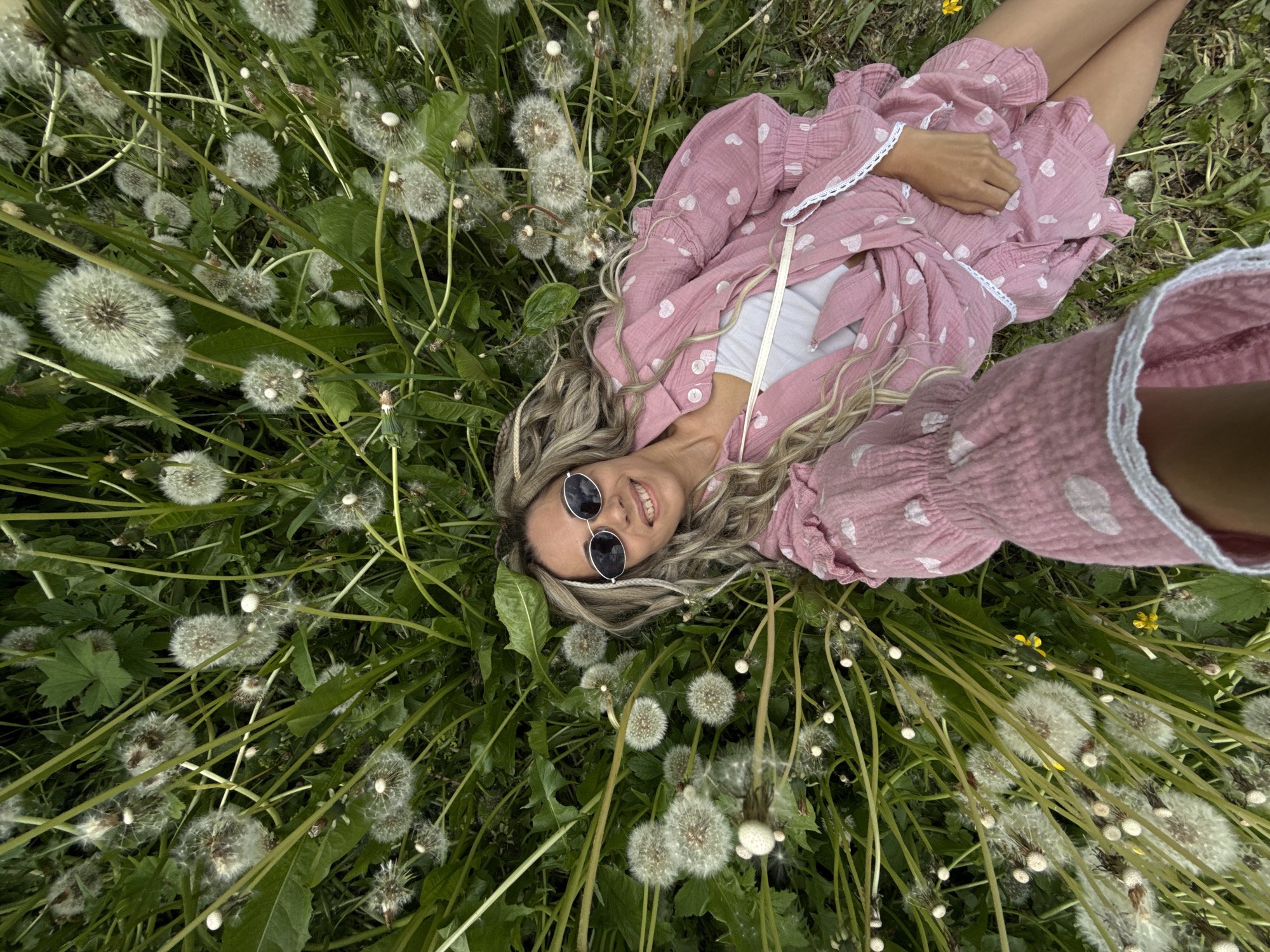 A young woman is lying on her back in a lush green field densely covered with white dandelion seed heads. The photograph is taken from a high angle, looking down at her, with her head positioned towards the lower center of the frame and her body extending diagonally upwards.
The woman has long, wavy blonde hair spread around her head on the grass. She is wearing a light pink, short-sleeved dress or robe adorned with small white heart patterns and white lace trim on the cuffs and edges. A white top is visible underneath her dress. She is also wearing round, dark-lensed sunglasses. Her bare legs are visible towards the upper left side of the image.
She has a relaxed and contented expression, with a gentle smile on her face as she looks up towards the camera. Her arms are casually placed, with one hand resting on her stomach. The setting is an outdoor natural environment, likely in Rusanovo, Russia, characterized by abundant dandelions and green foliage. The lighting appears soft and even, suggesting it is daytime with either an overcast sky or indirect sunlight. No text is visible in the image.
