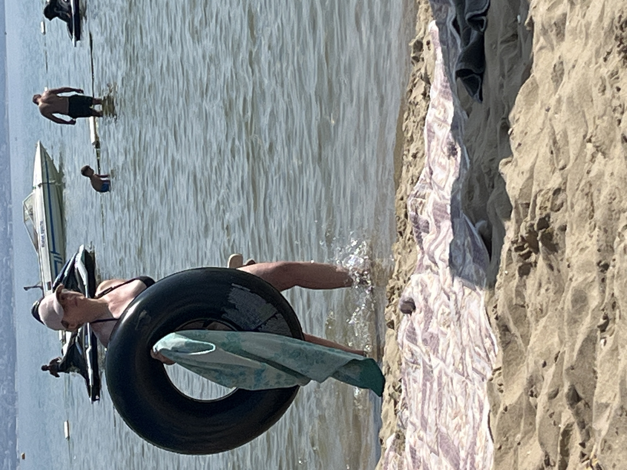 The image depicts a sunny day at a sandy beach with calm water. In the foreground, a woman wearing a bikini and a white cap is holding a large black inner tube. A light blue and white patterned towel is draped over the inner tube. The woman is partially submerged in the water, with her legs visible. Behind her, a white boat with blue accents is docked or anchored. Further in the background, a man in dark shorts stands in the water near a pole, and a young person in blue shorts is also in the water. The sand on the right side of the image is textured and uneven, with a few small rocks. In the distance, the shoreline with buildings can be seen, suggesting the location is Nesebar, Bulgaria. The overall impression is a relaxed scene at the beach during daytime, with people enjoying the water.