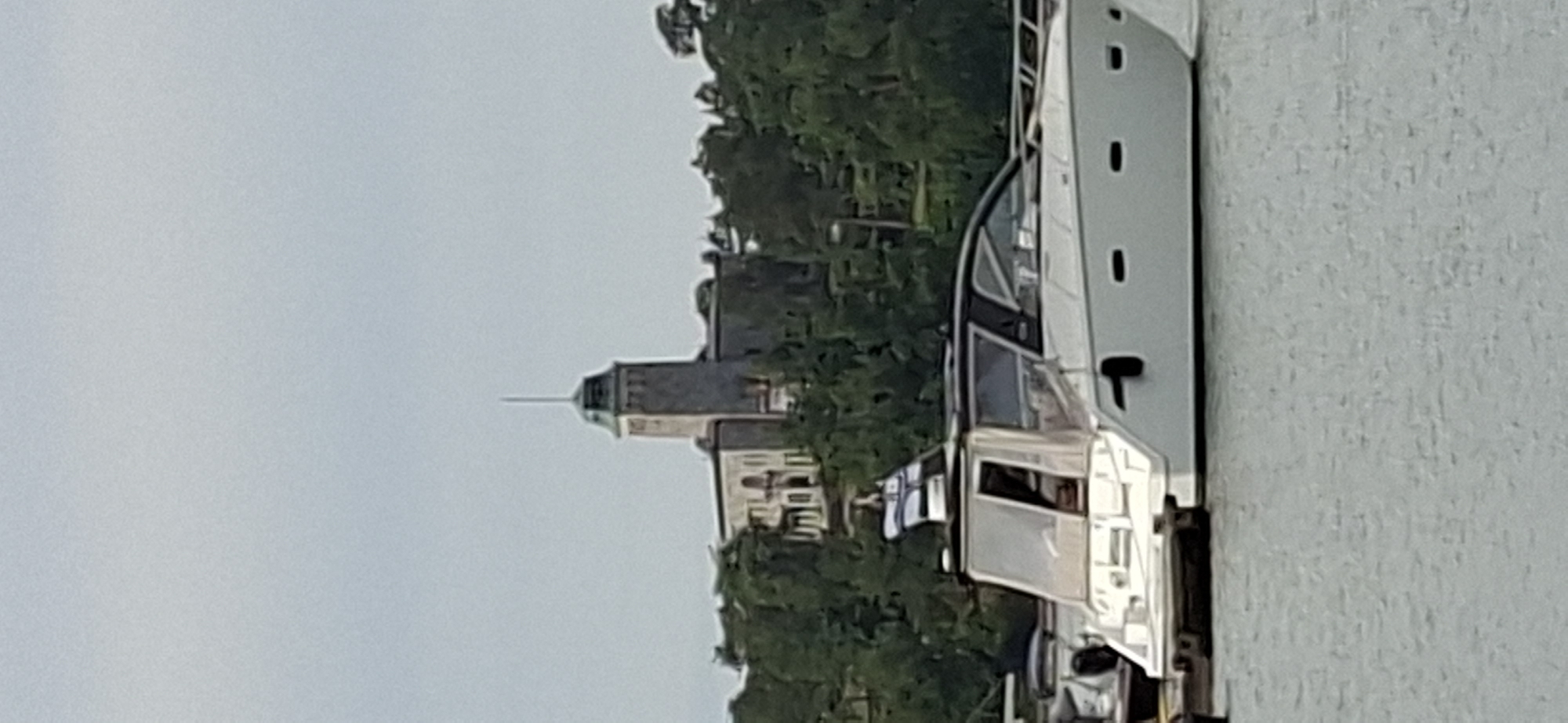 The image shows a white motor yacht docked in a body of water. In the background, on a hill surrounded by trees, is a distinctive stone building with a tall, square tower topped with a spire. The building appears to be a historical structure, possibly a castle or fortress. This structure is likely the main landmark of Naantali, Finland. The sky is overcast and the water is calm. There are no visible people in the image.