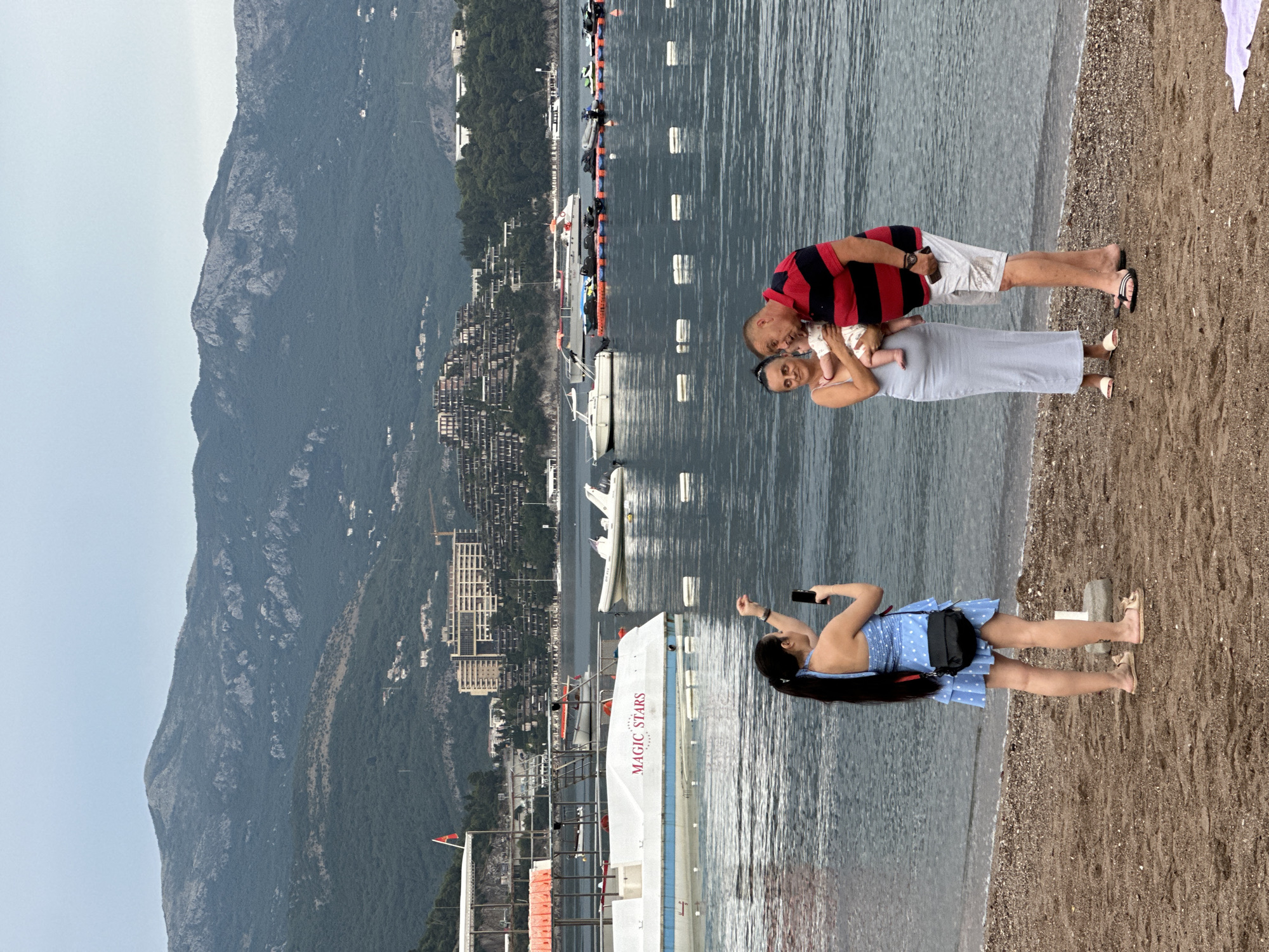 The image shows a family on a beach in Budva, Montenegro. A man in a red and black striped shirt and white shorts is holding a baby. A woman in a long, light blue dress is standing next to him. Another woman in a blue polka dot dress is on the sand taking a picture with her phone. In the background, there is a body of water with several boats, including a white boat with "MAGIC STARS" written on its side. Mountains covered in trees and buildings rise in the distance. The weather appears to be overcast, and the time of day is likely late afternoon or early evening, given the lighting. The overall mood seems relaxed and familial.