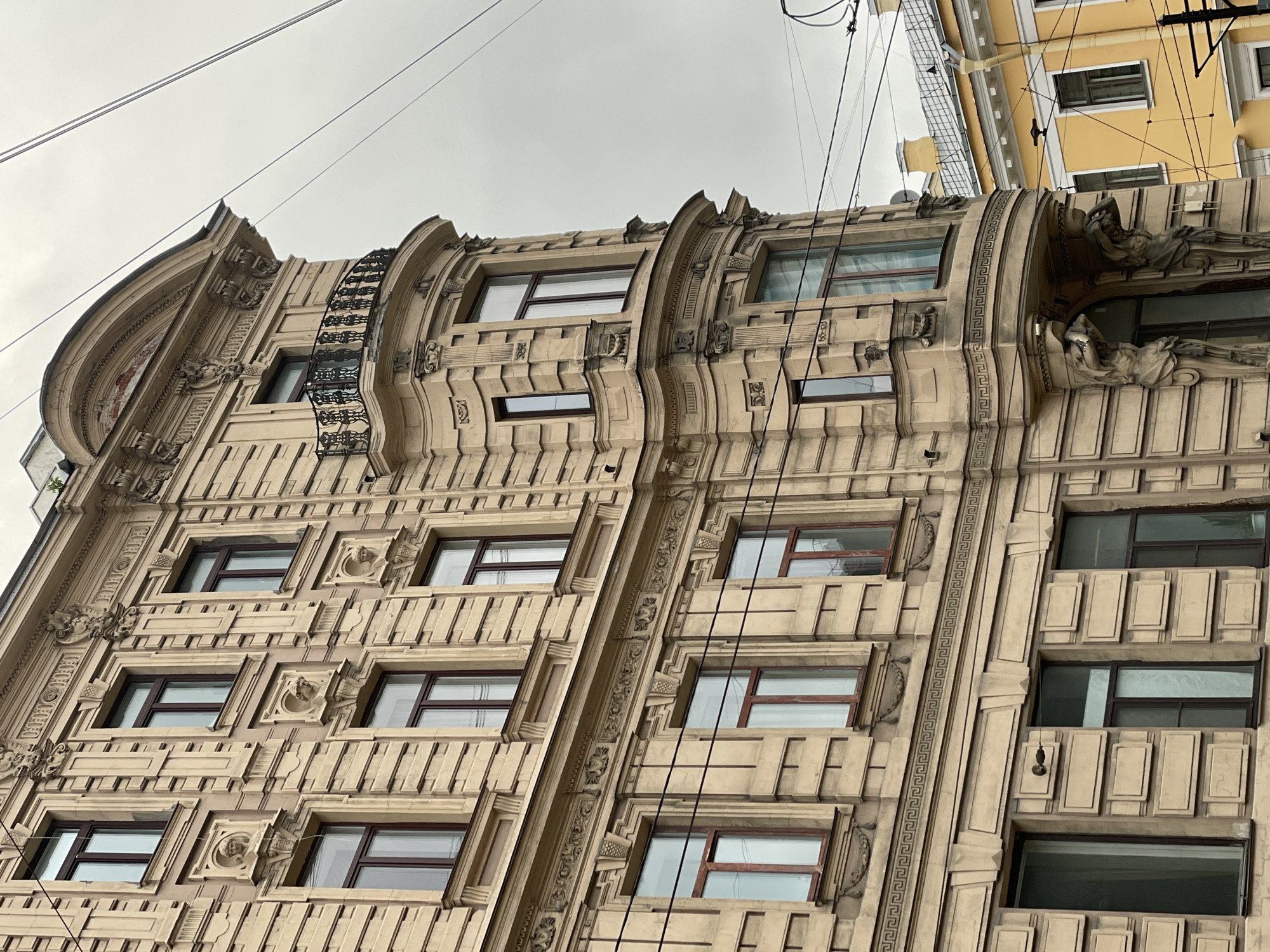 The image shows a detailed view of an ornate building facade in Saint Petersburg, Russia. The building has a light beige color and exhibits complex architectural details including decorative moldings, window frames, and classical motifs. Several windows are visible, recessed into the facade with dark frames. The building’s surface is textured with geometrical patterns. Power lines run across the sky in the background. Another yellow building is visible in the background. The sky is overcast and grey.