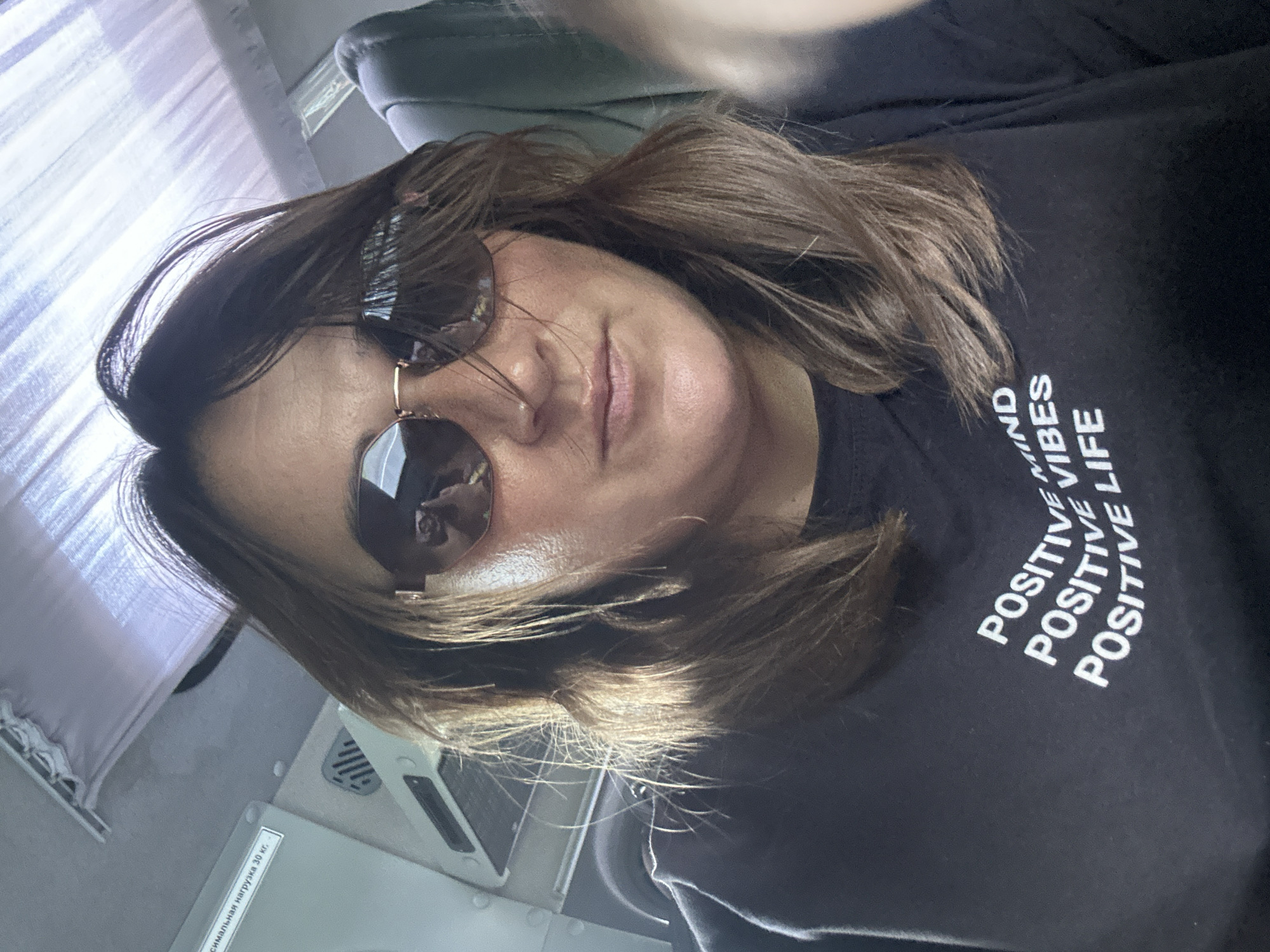 A woman is taking a selfie on what appears to be public transport. She is wearing sunglasses and a black t-shirt with white text that says "POSITIVE MIND POSITIVE VIBES POSITIVE LIFE." The background shows the interior of a vehicle, including seats and overhead compartments. There is also a sticker on the wall with Russian text that translates to "maximum load 30 kg."
