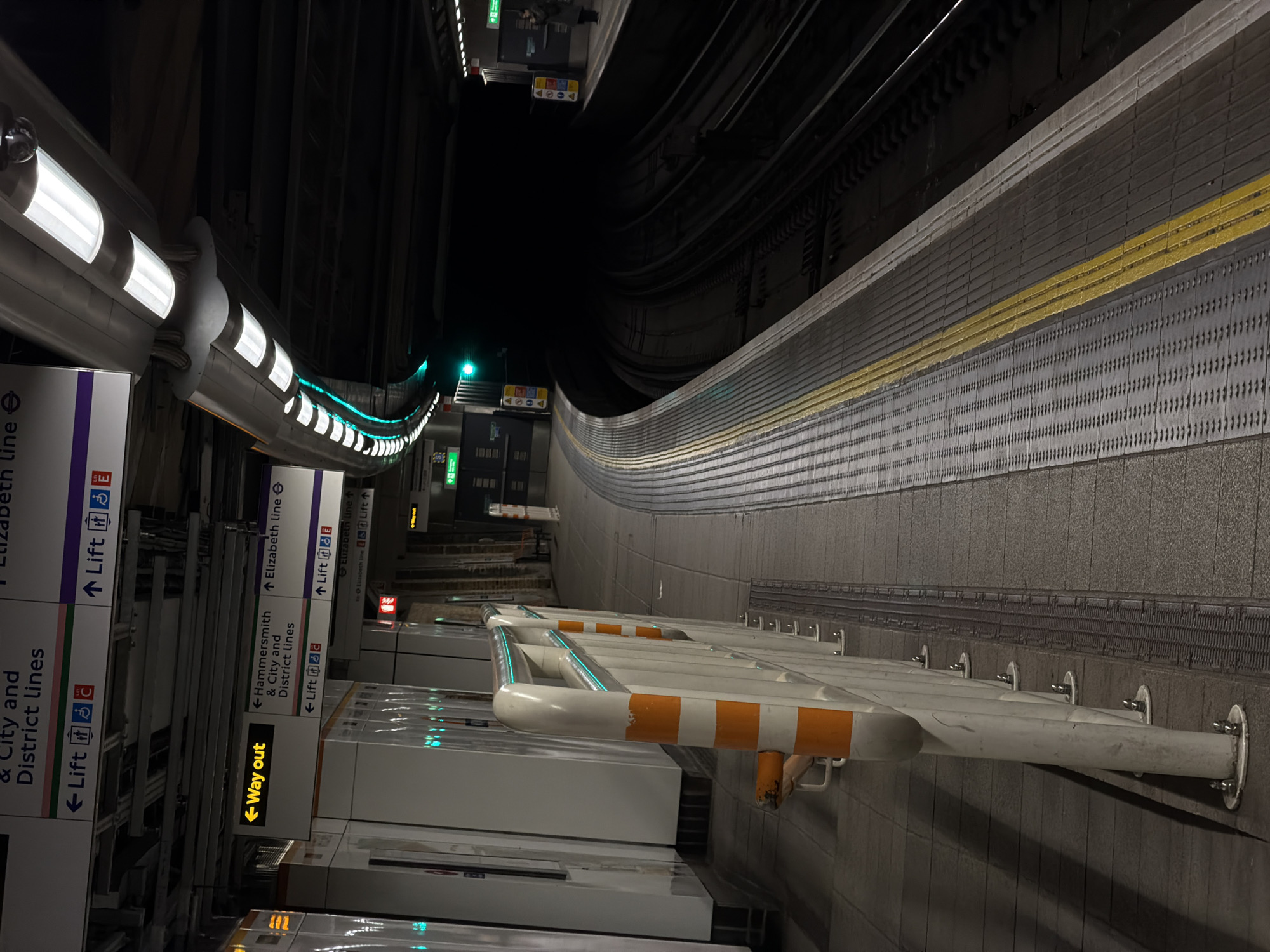 The image depicts a section of a London Underground station platform in the United Kingdom. The platform is illuminated by a series of white fluorescent lights arranged in a curved pattern along the ceiling. The platform edge is marked with a yellow line. The platform itself is grey and textured with tactile paving near the edge. A white metal barrier with orange markings runs along the platform. Signage indicates directions for the Elizabeth line, Hammersmith & City, and District lines, with arrows pointing towards lifts and "Way out" exits. The tunnel ahead is dark, with faint green lights visible in the distance, suggesting the direction of the track. The tracks themselves are visible above the platform edge. The platform curves, indicating the station is located on a bend in the line. The overall atmosphere is dim and utilitarian, typical of an underground station environment.