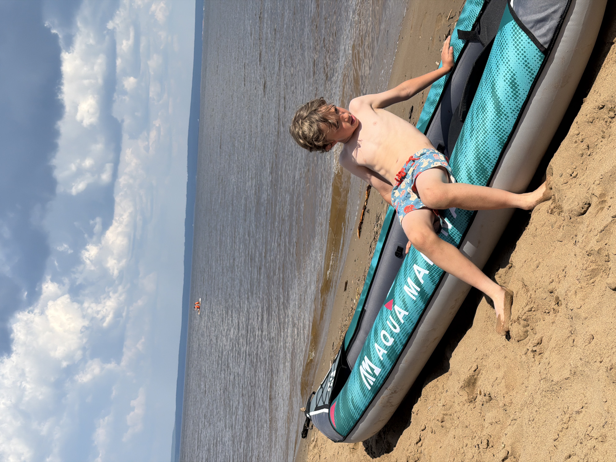 In Noviy, Russia, on a sandy beach, a young boy with fair hair sits on an inflatable kayak pulled ashore. The kayak is teal and gray, with the "Aqua Marina" logo visible. The boy wears patterned swim trunks and has sand on his feet. The water is calm and extends to the horizon under a partly cloudy sky. In the distance, on the water, two people on a white float are visible.