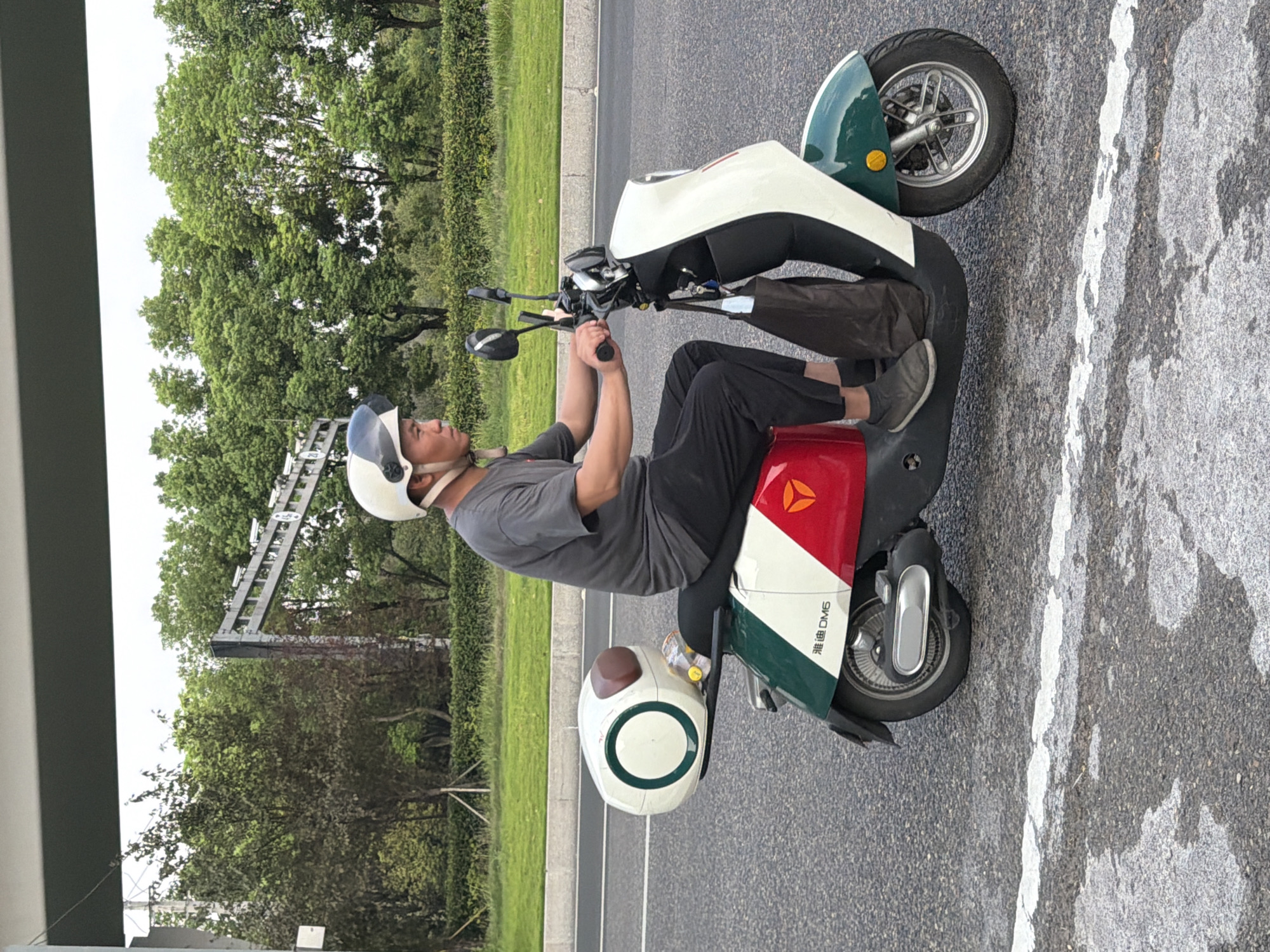 A man in a grey t-shirt, black pants, a white helmet with a clear visor, and grey sneakers is riding a white, green, and red electric scooter on an asphalt road with a white line marking. The scooter has a large white storage box on the back with a brown lid and a green ring design. A dark bag hangs from the handlebars. The man is looking forward, and his hands are on the handlebars. Behind him, a line of green trees and bushes is visible, with a grey overpass and traffic lights in the background. The image appears to be taken from the side, with the road in the foreground and the vegetation and infrastructure behind. The scene is consistent with an urban or suburban setting in Jiangjin, China.
