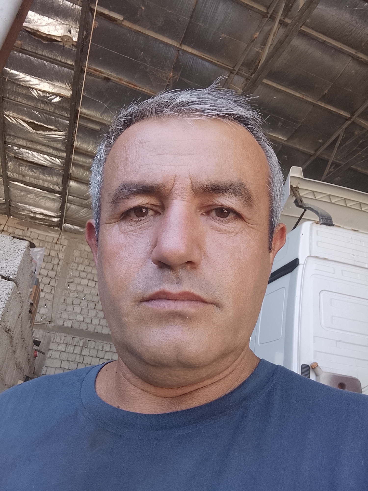 An eye-level, medium shot captures a man with graying hair, a dark blue t-shirt, and a tan complexion against an outdoor, industrial backdrop. The man is positioned in the foreground, with his face filling most of the frame. Behind him, a white truck cab is visible on the right, while the left side of the image shows a brick wall and a stack of white, porous materials. The overhead structure consists of exposed beams and insulation, suggesting an unfinished or utilitarian building. The lighting is bright, creating natural shadows on the man's face. The setting appears to be an outdoor storage or work area, possibly in a city like Sabir Rakhimov, Uzbekistan, given the presence of industrial elements.