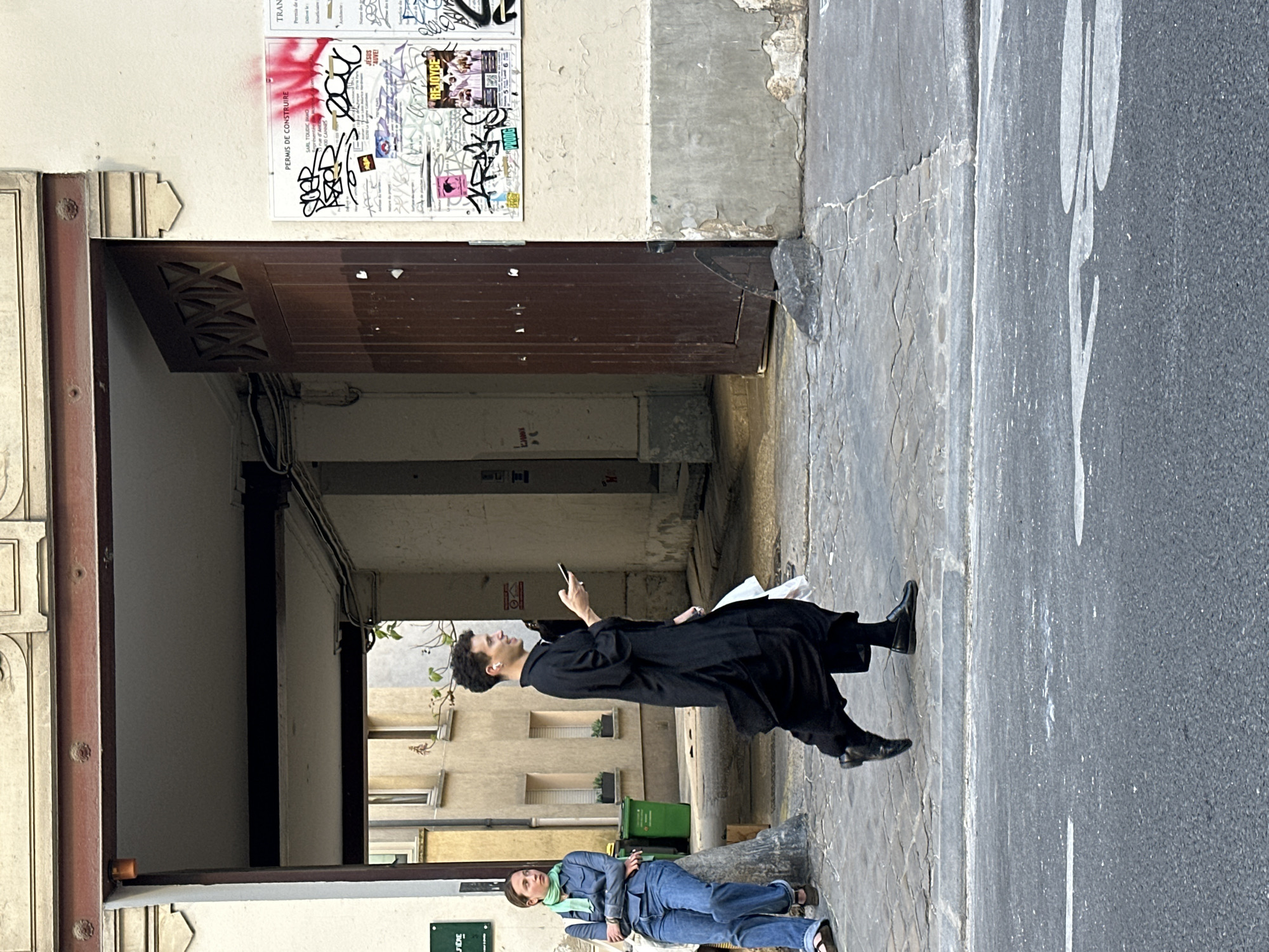 An outdoor, bright daylight scene captures a moment on a narrow urban street in Paris, featuring two individuals amidst old buildings and pavement.
On the left, an ancient-looking building facade with a prominent dark brown wooden archway entrance dominates the frame. The wall above this archway is densely covered with layered posters and various graffiti. Through the archway, glimpses of an inner courtyard with other building facades and a green public waste bin are visible. The ground consists of patterned cobblestone pavement on the left, transitioning to an asphalt road with a faded white marking on the right.
In the mid-ground, a person with dark, curly hair, dressed in a long black coat or tunic, black trousers, and black shoes, stands on the cobblestones. They are holding a white smartphone at eye level, appearing to be focused on its screen, and have a white earbud in their right ear. Closer to the foreground and slightly to the left, another person with light brown hair is visible. They are wearing a blue denim jacket, blue jeans, and a light green scarf, carrying a light-colored bag. This individual appears to be standing or leaning, looking towards the right side of the frame.
Notable text visible on the posters and graffiti includes "TRAN", "PERMIS DE CONSTRUIRE" (Building Permit), "SARL TOUDIC", "RODG", "JESUS AUVE!", "REJOYCE", and "FIERE". The scene portrays a casual, everyday street setting in a European city, likely Paris given the architectural cues and visible French text.