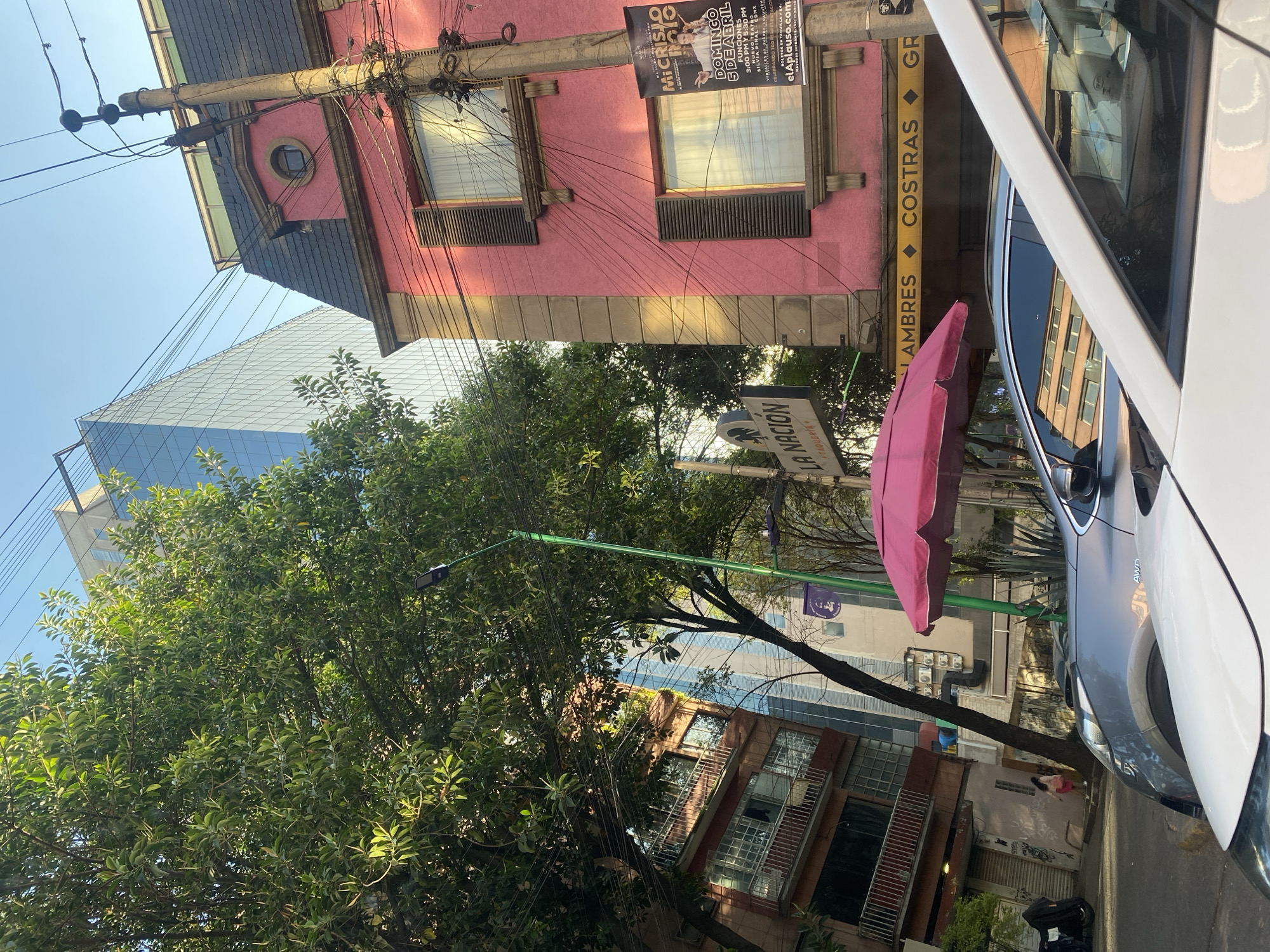 This upward-angled image captures a vibrant, sunlit urban street scene, likely in a Spanish-speaking country, characterized by a mix of architectural styles and abundant utility lines.
In the foreground, a partial view of a white car, possibly an SUV, occupies the right side, with a darker grey SUV visible next to it. A bright pink outdoor umbrella stands out in the lower mid-ground. Above the street, a green light pole extends horizontally.
The central feature is a charming two-story building painted a distinct pink, topped with a dark, possibly tiled, roof. Its facade displays several windows, some with louvered shutters, and a unique circular window on its upper level. A striking element is the dense network of black electrical wires crisscrossing the building's exterior and an adjacent utility pole. A poster affixed to the pink building advertises "MI CRISIS ROJO" (My Red Crisis), mentioning "DOMINGO 5 DE ABRIL", "FUNCIONES 3:00 PM Y 5:30 PM", and "elAplauso.com" for tickets. Other text on the building or signs includes "LAMBRES", "COSTRAS", and "GR".
Behind the pink building and lush green trees, a modern glass skyscraper with a grid pattern reflects the clear blue sky, providing a stark contrast between old and new architecture. Further down the street, obscured by foliage, another building with brick or brown siding and glass block windows is visible. A sign for "LA NACION" and "TAQUERI" can be seen amidst the trees. In the distance, a person in a pink top and white pants is visible between buildings.
The bright light and clear blue sky indicate daytime. The scene conveys a busy, somewhat cluttered, yet lively urban environment.