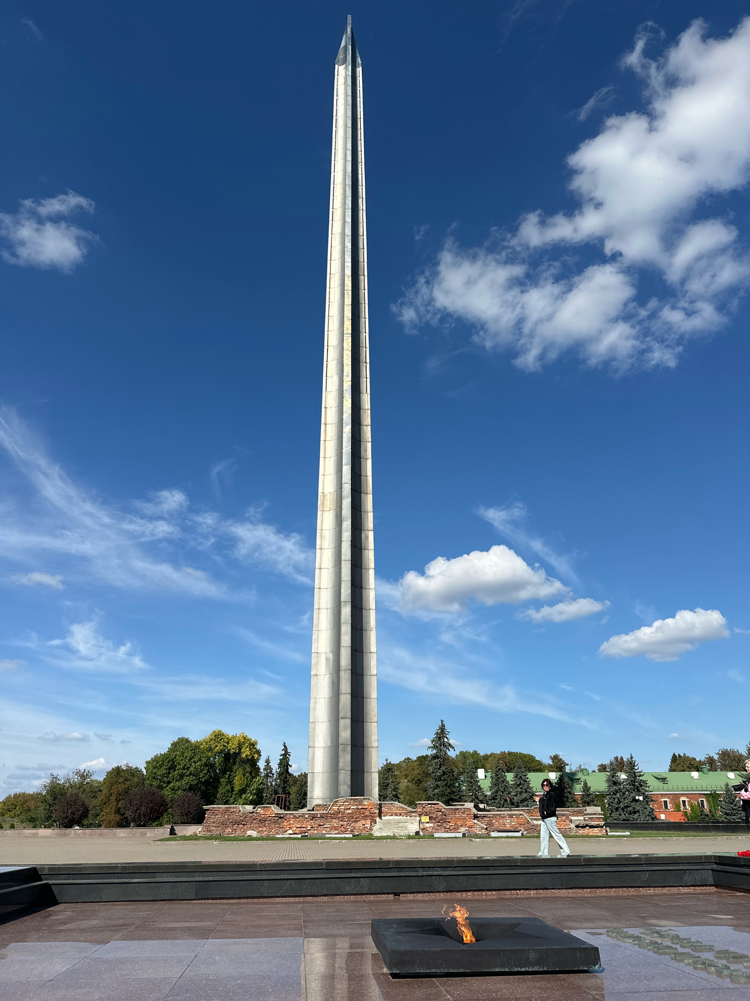 In Brest, Belarus, the Monument of Courage, a soaring obelisk of concrete and metal, dominates the frame against a clear blue sky dotted with white clouds. The monument, a slender, angular spire, stands tall and imposing. At its base, a low, rough-hewn brick wall hints at historical ruins. In the foreground, a dark, square structure with an eternal flame burning within it serves as a focal point. To the right of the monument, a person in a black jacket and light-colored pants walks casually, silhouetted against the background. Behind the monument and the brick wall, a row of evergreen trees and deciduous trees form a verdant backdrop, leading to red-brick buildings with green roofs. The scene is set in an open, paved area, suggesting a memorial or park.