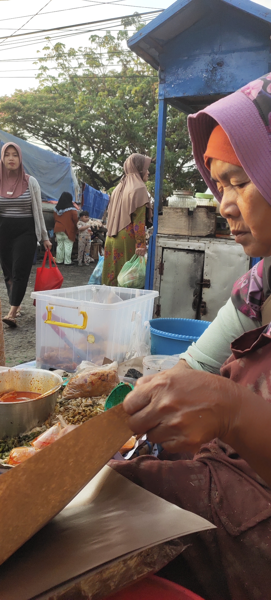 The image captures a scene at a street food stall in Karangtowo, Indonesia. In the foreground, a woman wearing a purple headscarf and an orange underscarf is in the process of preparing food. She is holding a piece of brown paper, and her hands are visible as she works. Various food ingredients and containers are spread out on a table in front of her. To the left, a silver bowl contains a reddish sauce or soup. Behind this, a clear plastic container with yellow handles holds more food items. To the right, a blue basin is visible. In the mid-ground, several other people are present. A woman in a light brown headscarf and a green patterned dress is holding a green plastic bag. Another woman in a pink headscarf and a grey cardigan is walking away from the camera, carrying a red bag. A young child in a patterned outfit stands between them. The background shows trees and a blue canopy structure, characteristic of an outdoor market or roadside vendor setup. The overall atmosphere appears to be that of a bustling marketplace during daylight.