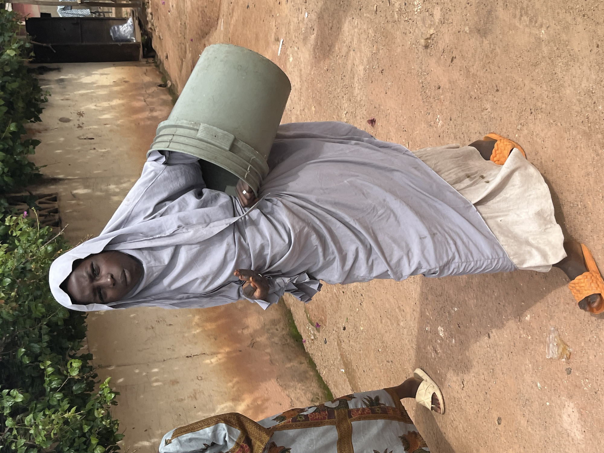A young person wearing a light grey hijab and matching flowing garments is carrying a large, grey plastic bucket in the city of Jos, Nigeria. The person is also wearing bright orange slides on their feet and a ring on their left hand. Another person, partially visible on the left, wears a patterned blue and gold traditional outfit and cream-colored slides. The background consists of a textured, light-colored wall with some staining and green foliage growing above it. The ground is a dusty, reddish-brown color, and there are small pieces of debris scattered around. The overall scene suggests an outdoor environment, possibly a street or courtyard.