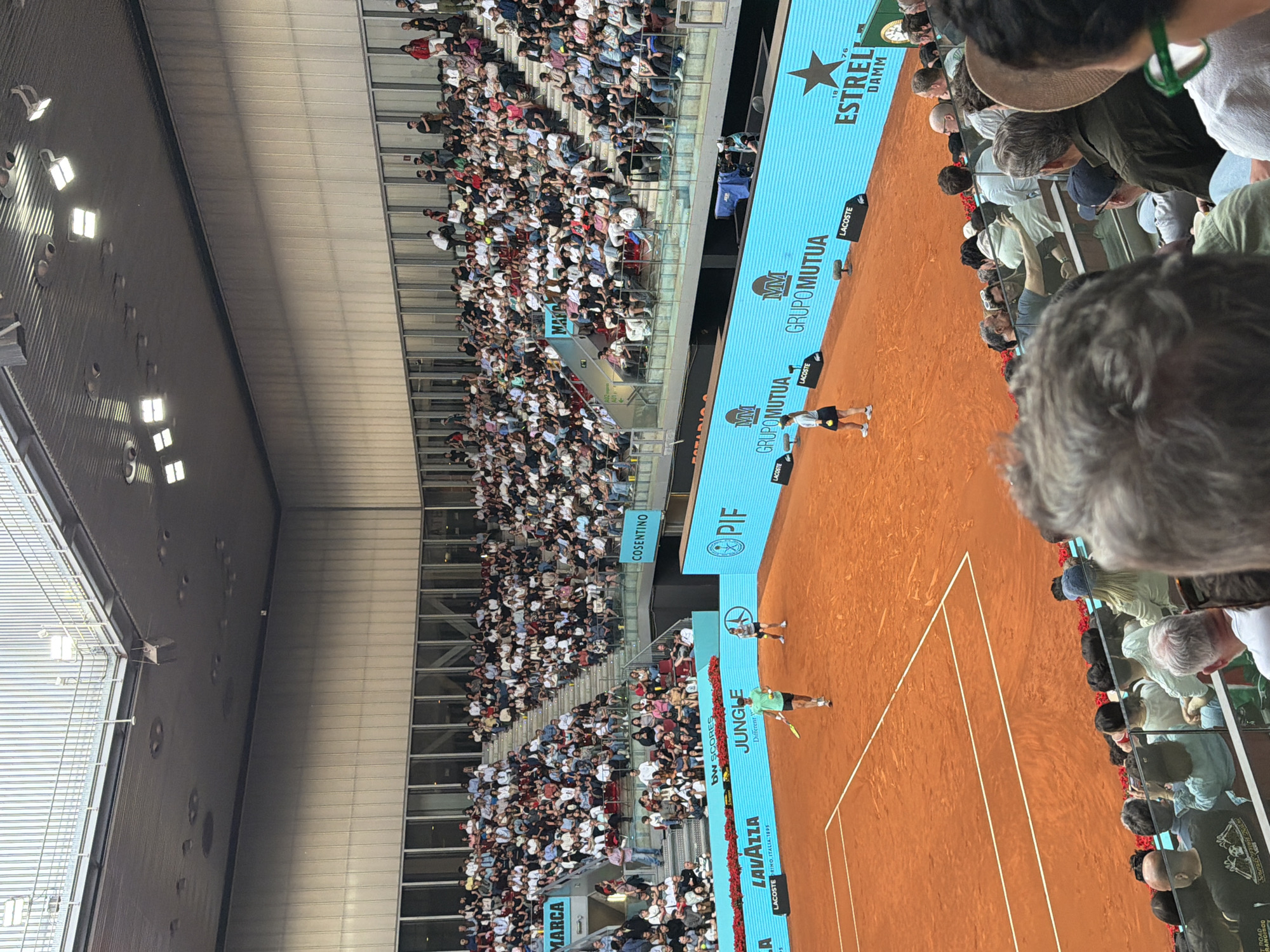 The image shows a tennis match being played on a clay court in an arena in Madrid, Spain. Two players are visible on the court. The stands are filled with spectators. Numerous advertisements are displayed around the court. The environment includes a stadium roof and lighting fixtures. The scene appears to be captured from a high vantage point within the arena.